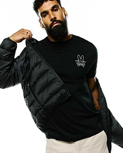 Black Mens Emerik Hooded Puffer Jacket with a quilted design, worn over a classic fit black jersey t-shirt featuring a prominent white Psycho Bunny skull logo on the left chest, paired with beige pants.