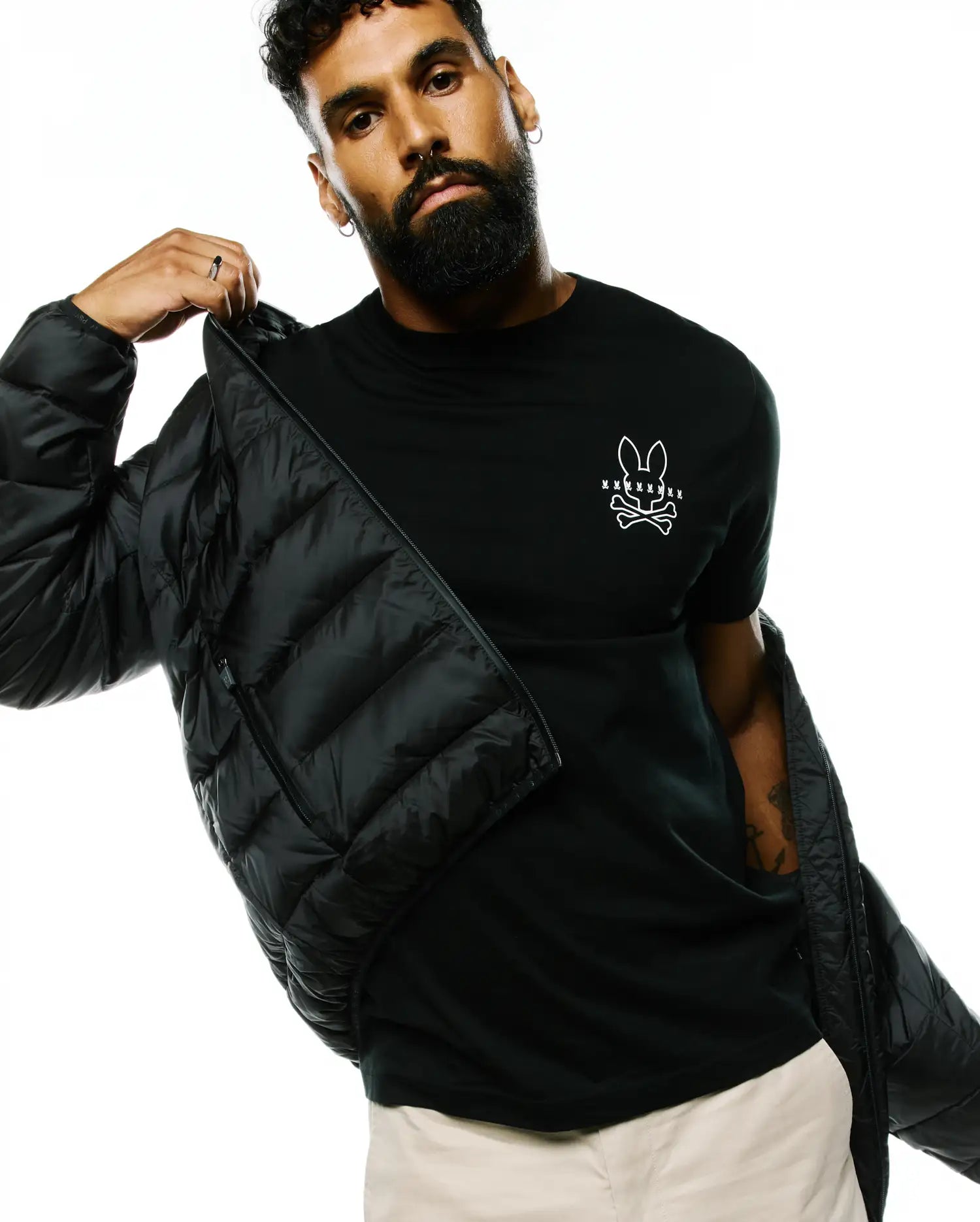 Black Mens Emerik Hooded Puffer Jacket with a quilted design, worn over a classic fit black jersey t-shirt featuring a prominent white Psycho Bunny skull logo on the left chest, paired with beige pants.