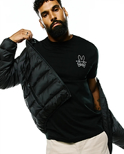 Black Mens Emerik Hooded Puffer Jacket with a quilted design, worn over a classic fit black jersey t-shirt featuring a prominent white Psycho Bunny skull logo on the left chest, paired with beige pants.