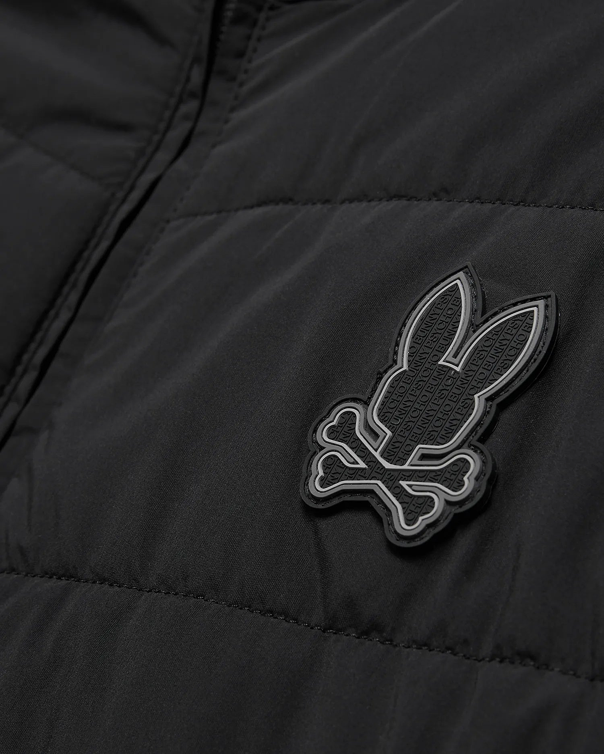 Mens Gordley Mix Media Jacket, black quilted outerwear with a prominent grey and white Psycho Bunny skull logo on the chest, featuring subtle repeating brand text within its design.