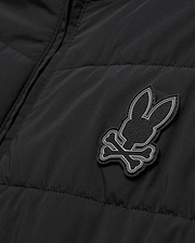 Mens Gordley Mix Media Jacket, black quilted outerwear with a prominent grey and white Psycho Bunny skull logo on the chest, featuring subtle repeating brand text within its design.