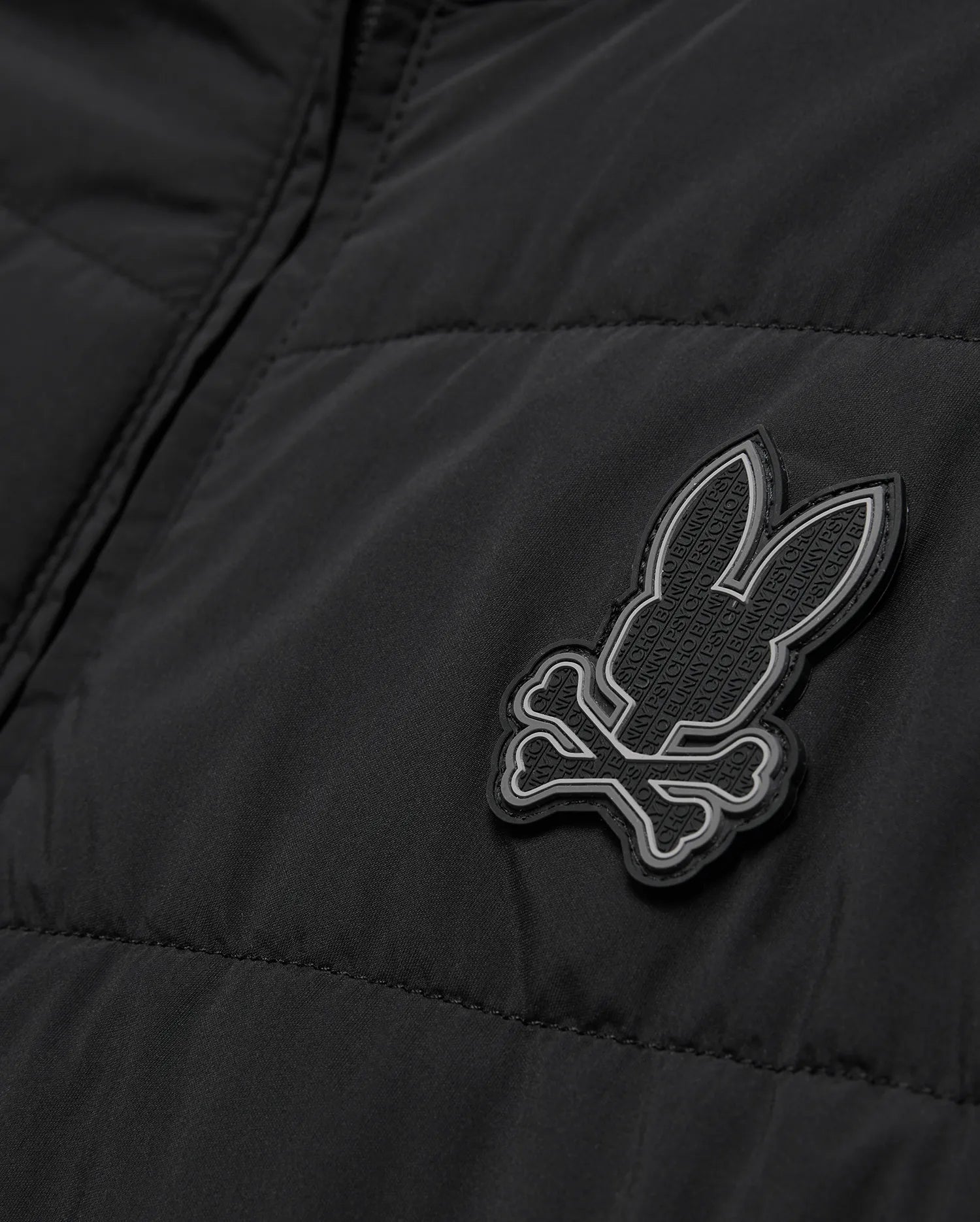 Mens Gordley Mix Media Jacket, black quilted outerwear with a prominent grey and white Psycho Bunny skull logo on the chest, featuring subtle repeating brand text within its design.