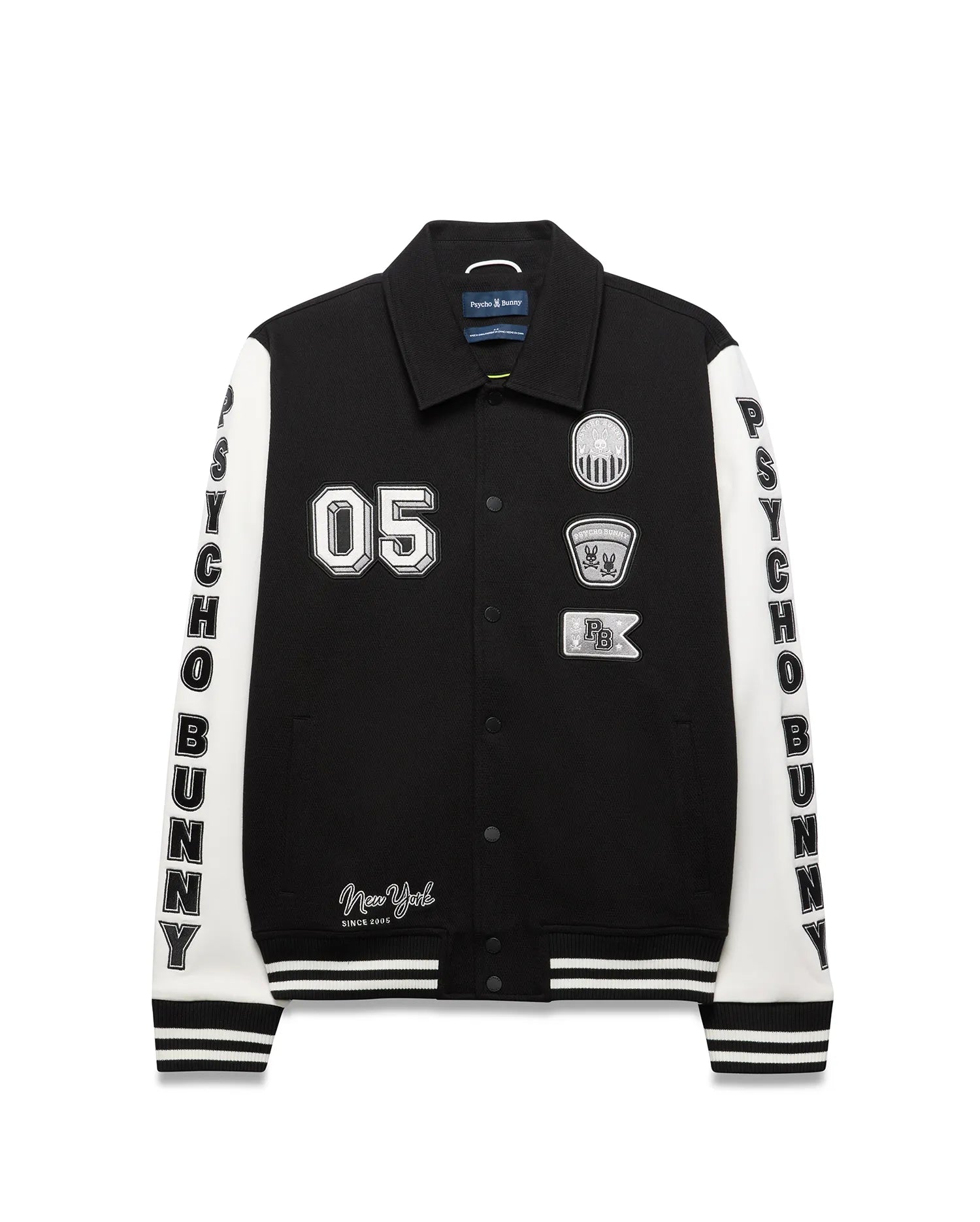 Mens Reilly Knit Coach Jacket, black with white sleeves, features embroidered 05, New York text, multiple Psycho Bunny skull logo patches, and vertical PSYCHO BUNNY text on sleeves, with striped cuffs and hem. (5)