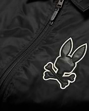 Mens Nathaniel Satin Coach Jacket in black, featuring a prominent embroidered Psycho Bunny skull logo on the chest with black leather-like texture and a white outline, alongside a black zipper. (6)
