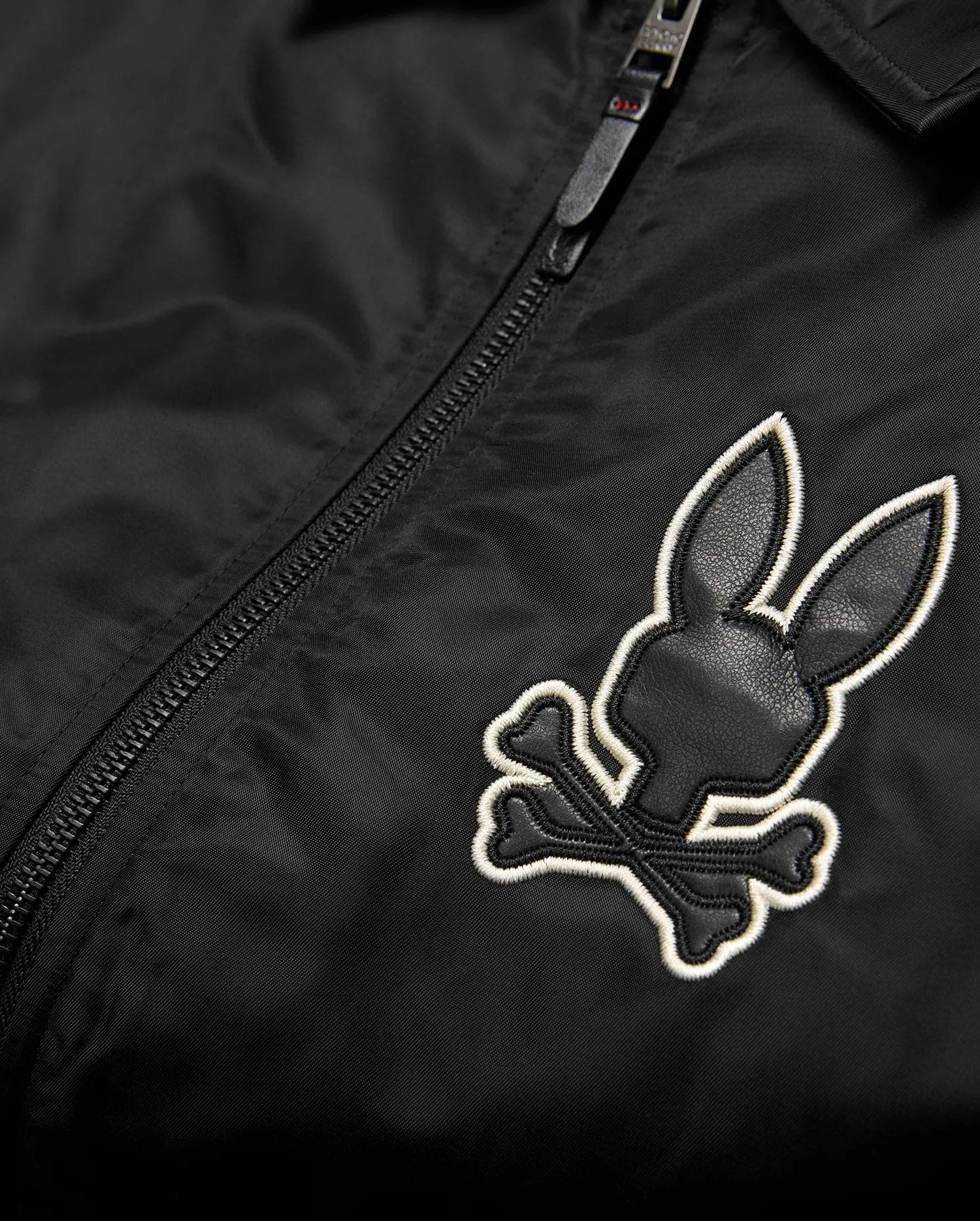 Mens Nathaniel Satin Coach Jacket in black, featuring a prominent embroidered Psycho Bunny skull logo on the chest with black leather-like texture and a white outline, alongside a black zipper. (6)