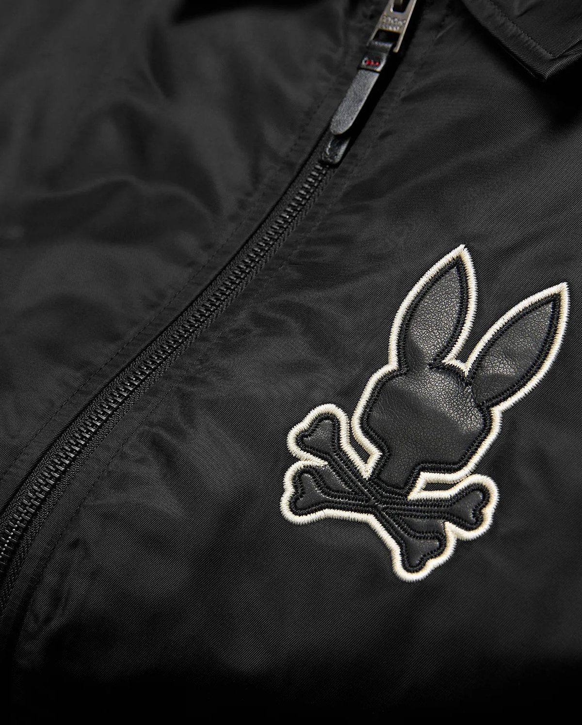 Mens Nathaniel Satin Coach Jacket in black, featuring a prominent embroidered Psycho Bunny skull logo on the chest with black leather-like texture and a white outline, alongside a black zipper. (6)