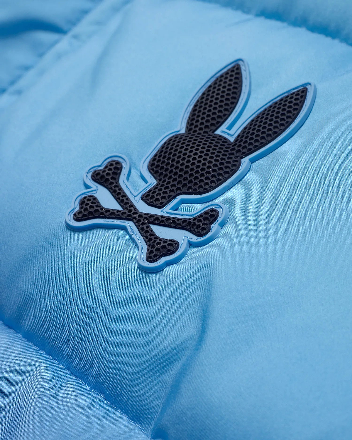 Men's Solan Puffer Jacket in sky blue, featuring a smooth, padded nylon texture and a rubberized dark grey Psycho Bunny skull logo with a light blue outline on the chest. (6)