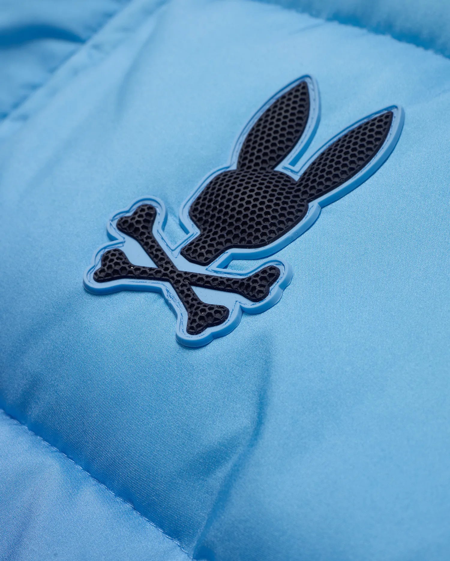 Men's Solan Puffer Jacket in sky blue, featuring a smooth, padded nylon texture and a rubberized dark grey Psycho Bunny skull logo with a light blue outline on the chest. (6)