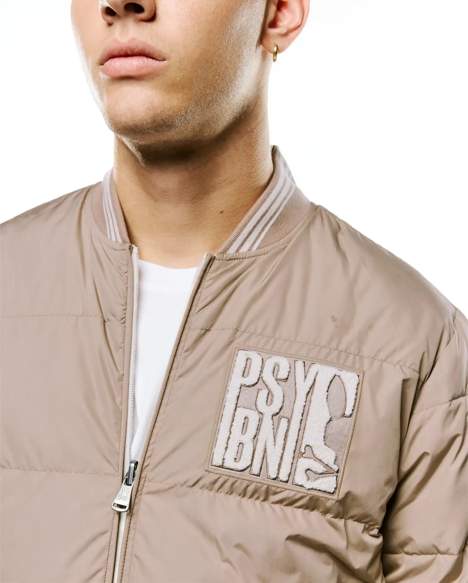 Men's Franco Reversible Bomber in khaki padded nylon, featuring a ribbed collar with white stripes, a large textured cream PSY BN and bunny skull patch on the left chest, and a bunny skull zipper pull. (3)
