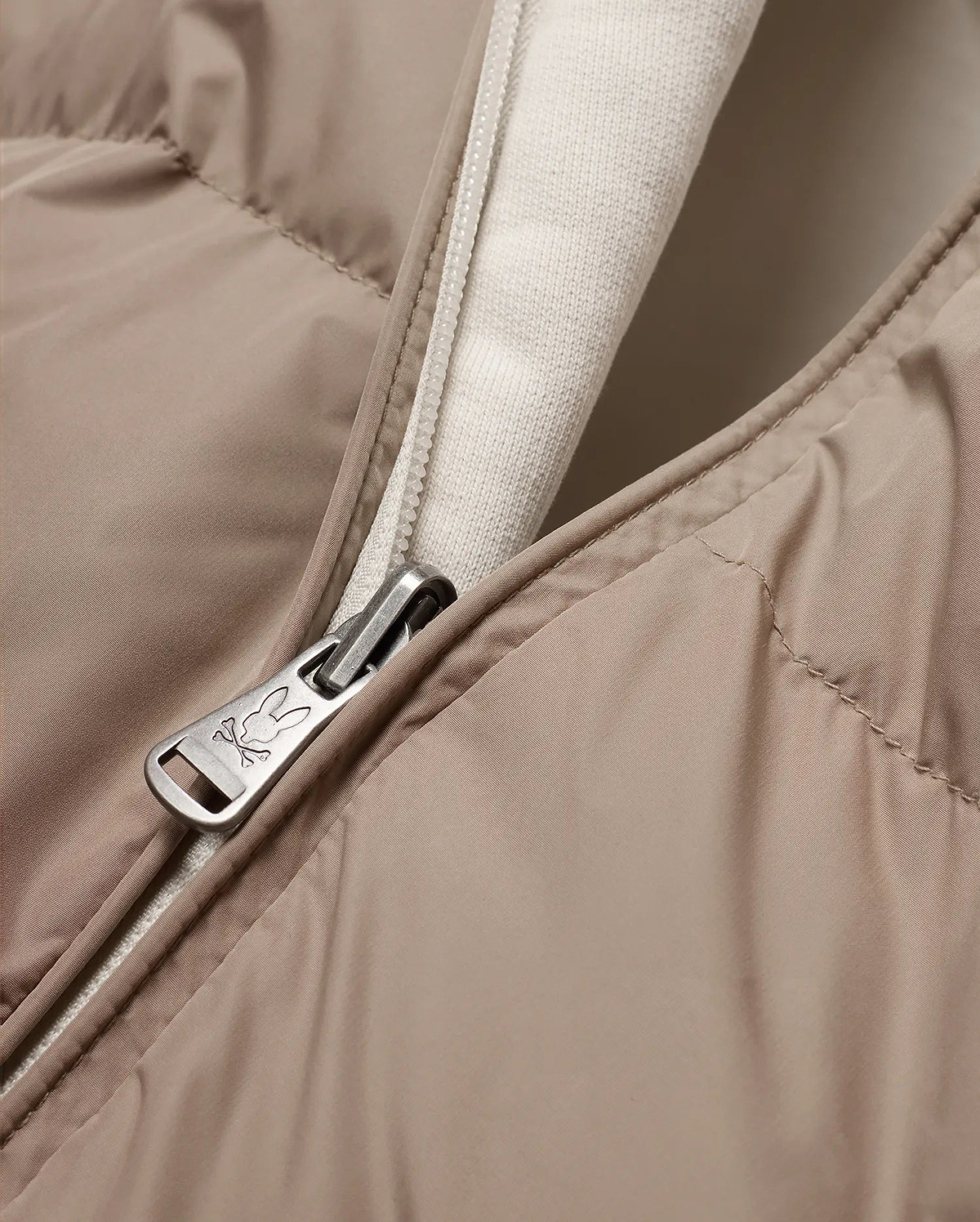 Men's Franco Reversible Bomber, a beige padded jacket with a cream knit lining and a full-zip closure featuring a silver Psycho Bunny skull logo engraved zipper pull. (6)