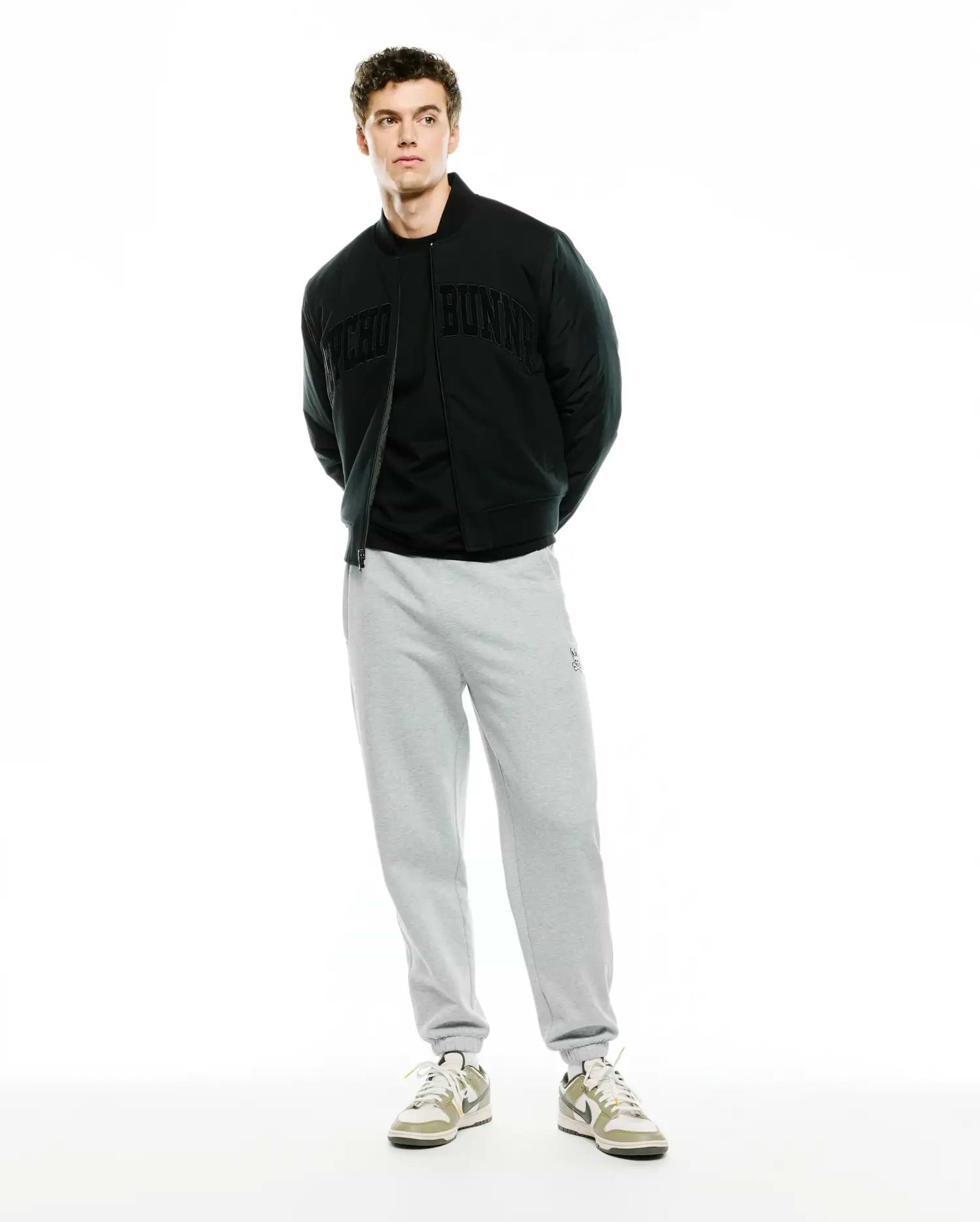 Mens Joaquin Mix Media Reversible Bomber in black, featuring large tonal Psycho Bunny lettering across the chest, paired with light grey relaxed fit french terry sweatpants with a small embroidered bunny skull logo on the left thigh.