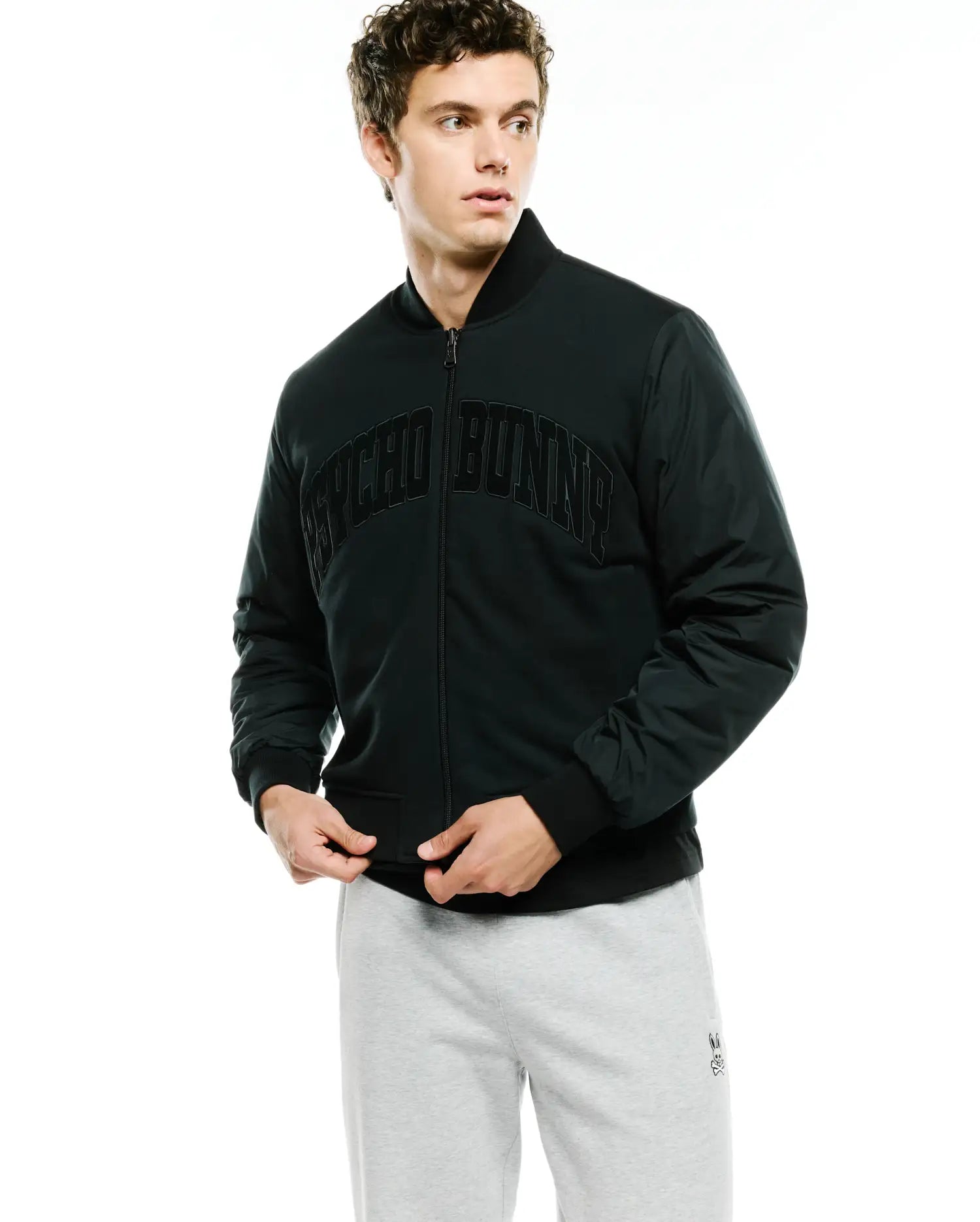 Mens Joaquin Mix Media Reversible Bomber in dark green, featuring a classic fit, full zip, ribbed black collar, cuffs, and hem, with a large arched black PSYCHO BUNNY text logo across the chest and contrasting sleeve fabric.