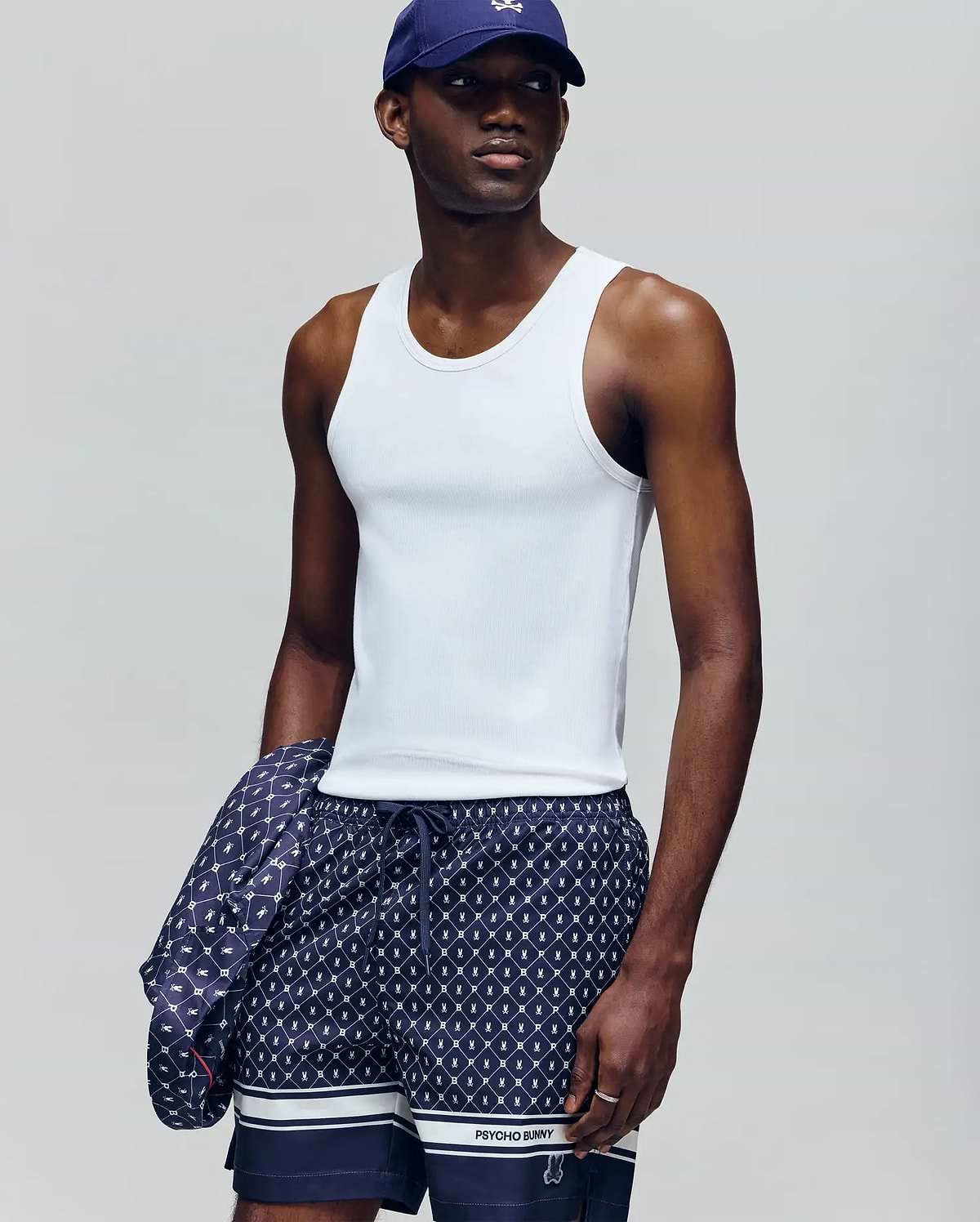A man wears a Psycho Bunny MENS 2 PACK TANK TOP - MO1400001 in white, navy patterned shorts, and a navy cap, holding a matching jacket. He stands against a plain light gray background, looking slightly to the side.