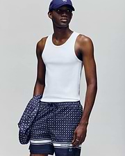 A man wears a Psycho Bunny MENS 2 PACK TANK TOP - MO1400001 in white, navy patterned shorts, and a navy cap, holding a matching jacket. He stands against a plain light gray background, looking slightly to the side.
