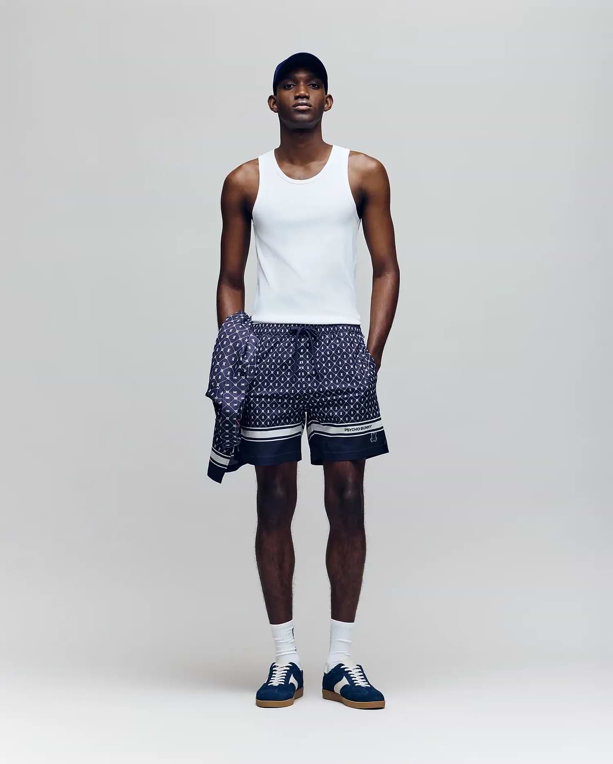 A man wears a Psycho Bunny MENS 2 PACK TANK TOP - MO1400001 in white, with patterned shorts, sneakers, white socks, a dark baseball cap, and a matching patterned shirt tied around his waist against a plain background.
