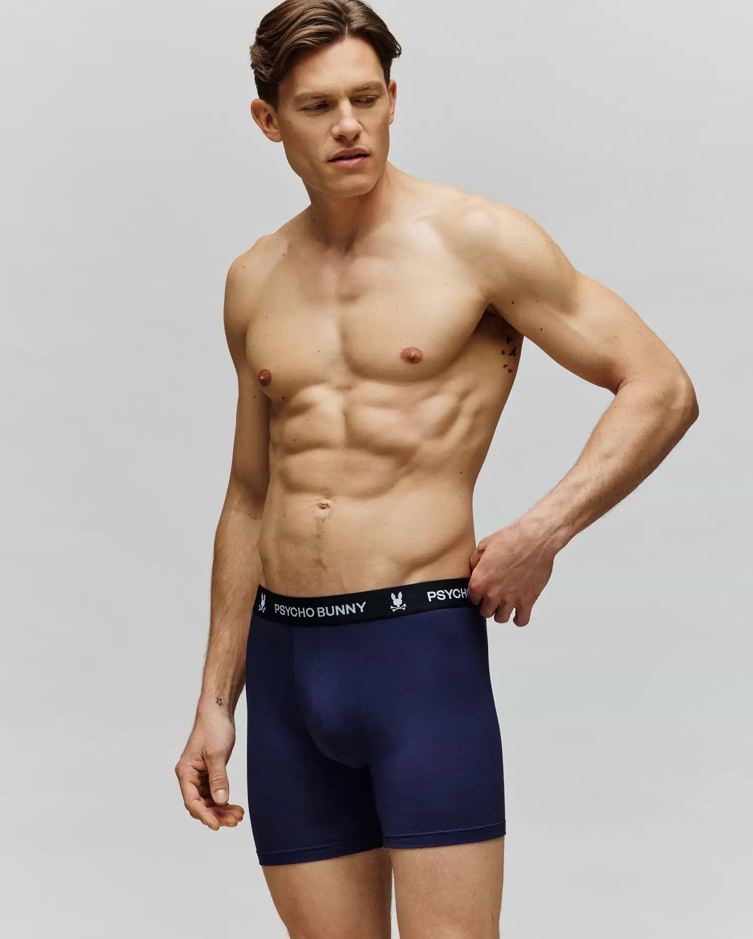 A muscular man models the Psycho Bunny MENS 3 PACK BOXER BRIEF - MO1400006, featuring navy blue cotton-modal boxers with a black "Psycho Bunny" waistband. He stands against a plain background, gazing left with one hand on his waistband.