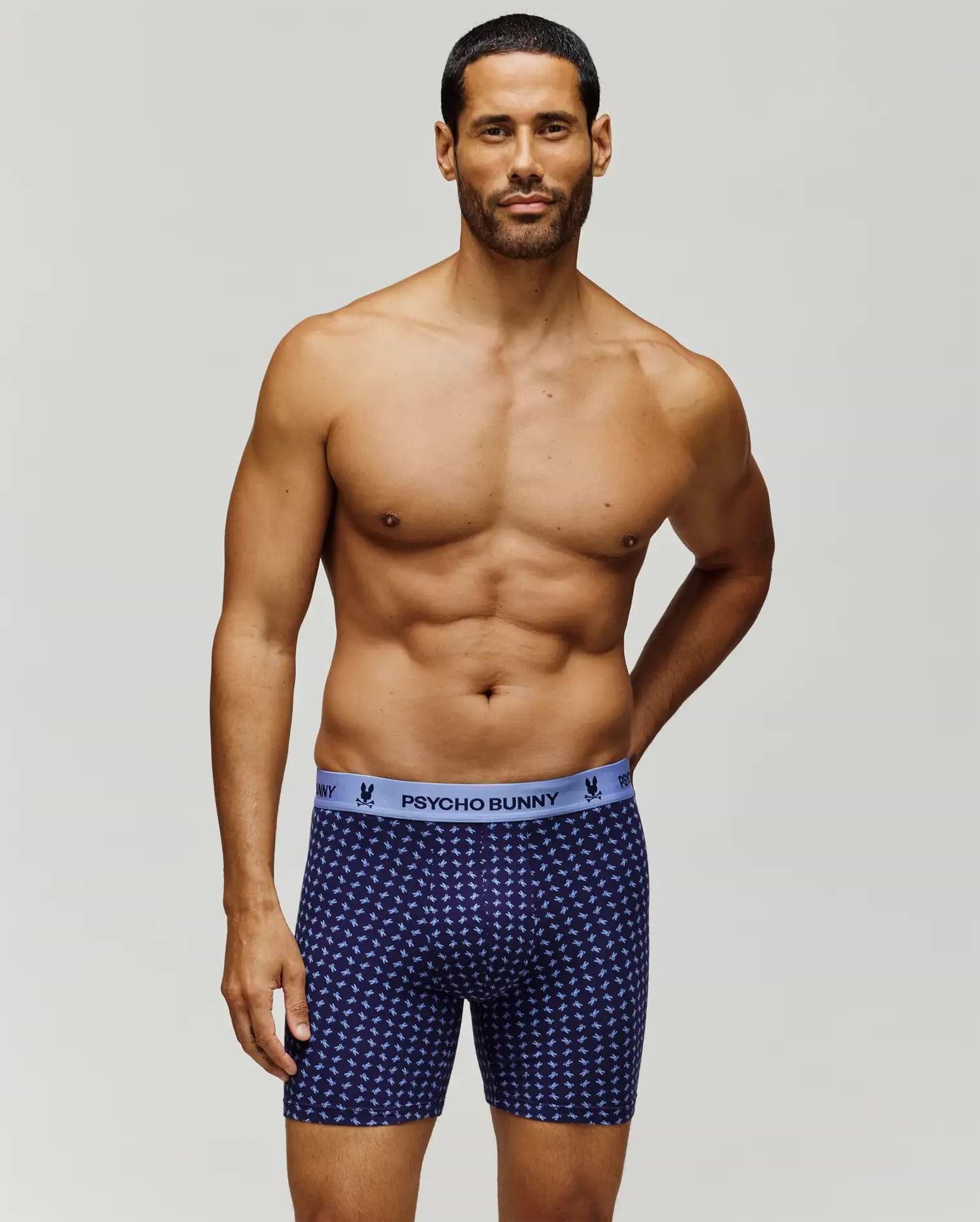 A muscular man with short dark hair stands against a plain background, wearing Psycho Bunny's MENS SINGLE BOXER BRIEF - MO1400010 in patterned navy blue Pima cotton with a light blue "Psycho Bunny" waistband for all-day comfort. (1)