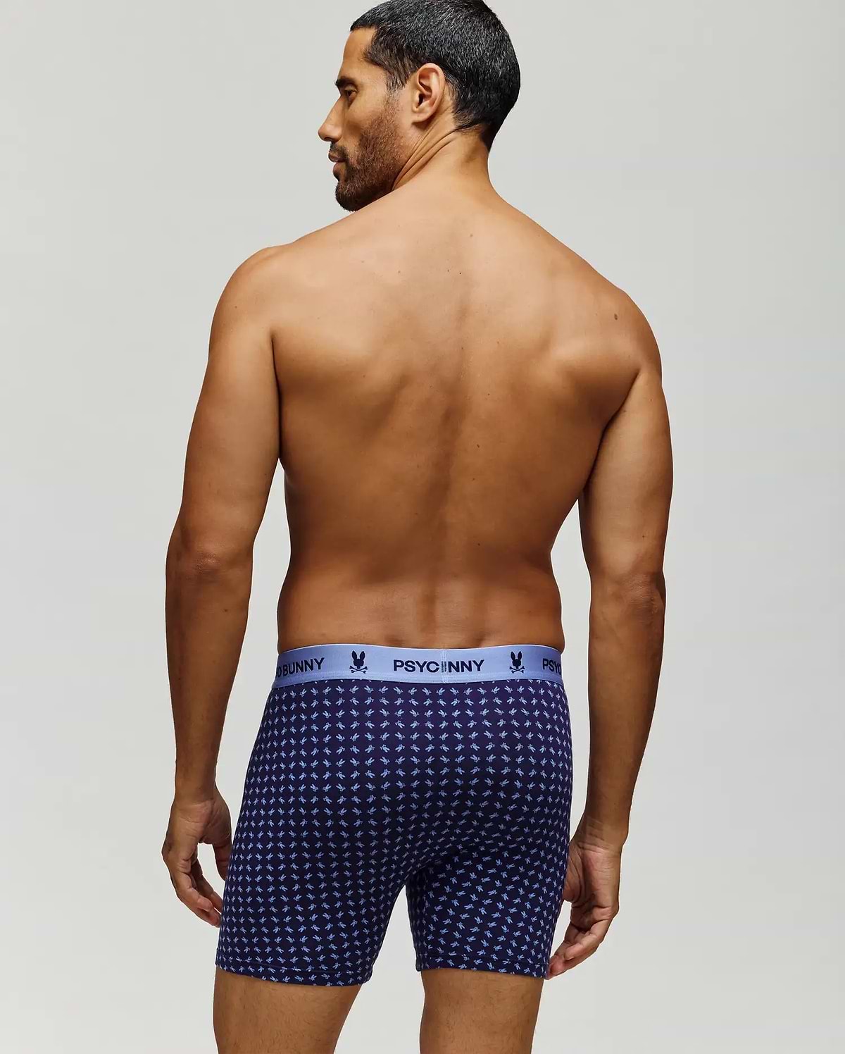 A man with short dark hair stands shirtless, facing away, wearing navy blue Psycho Bunny MENS SINGLE BOXER BRIEF - MO1400010 with a light blue logo waistband and breathable knit Pima cotton for comfort and style. (3)
