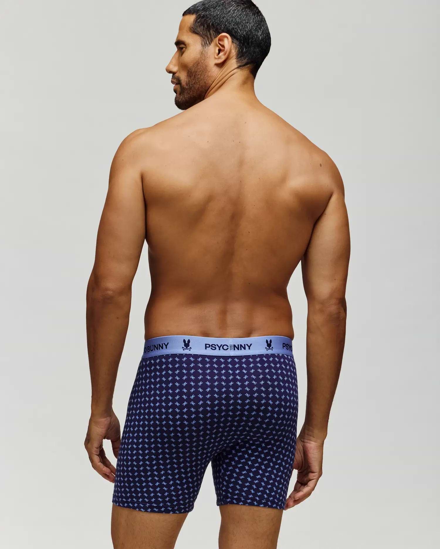 A man with short dark hair stands shirtless, facing away, wearing navy blue Psycho Bunny MENS SINGLE BOXER BRIEF - MO1400010 with a light blue logo waistband and breathable knit Pima cotton for comfort and style. (3)