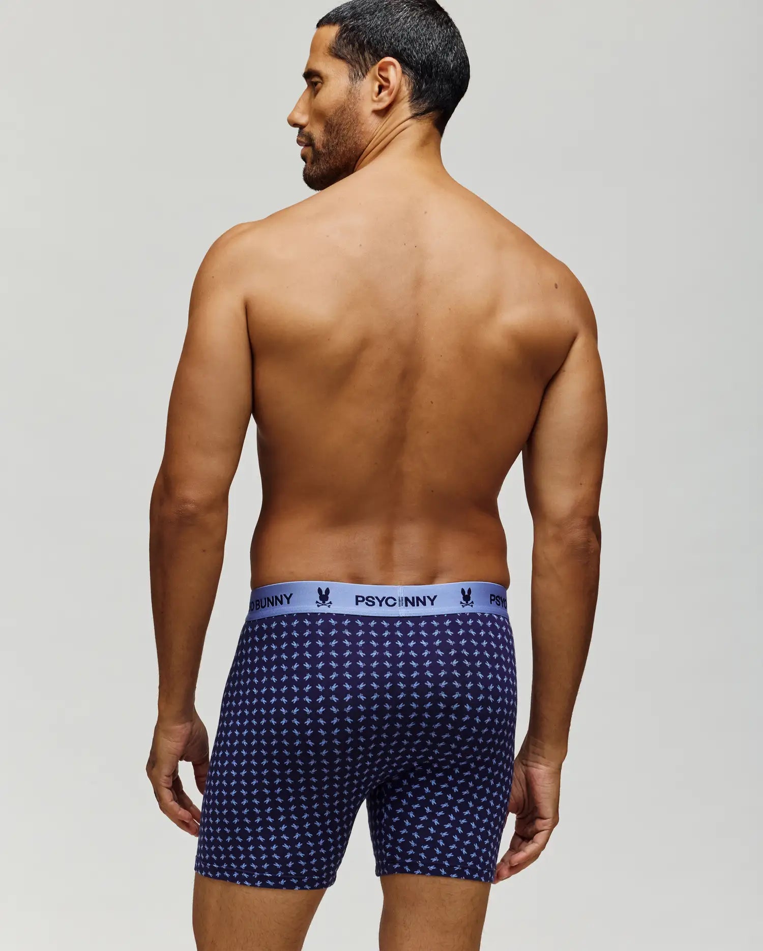 A man with short dark hair stands shirtless, facing away, wearing navy blue Psycho Bunny MENS SINGLE BOXER BRIEF - MO1400010 with a light blue logo waistband and breathable knit Pima cotton for comfort and style. (3)
