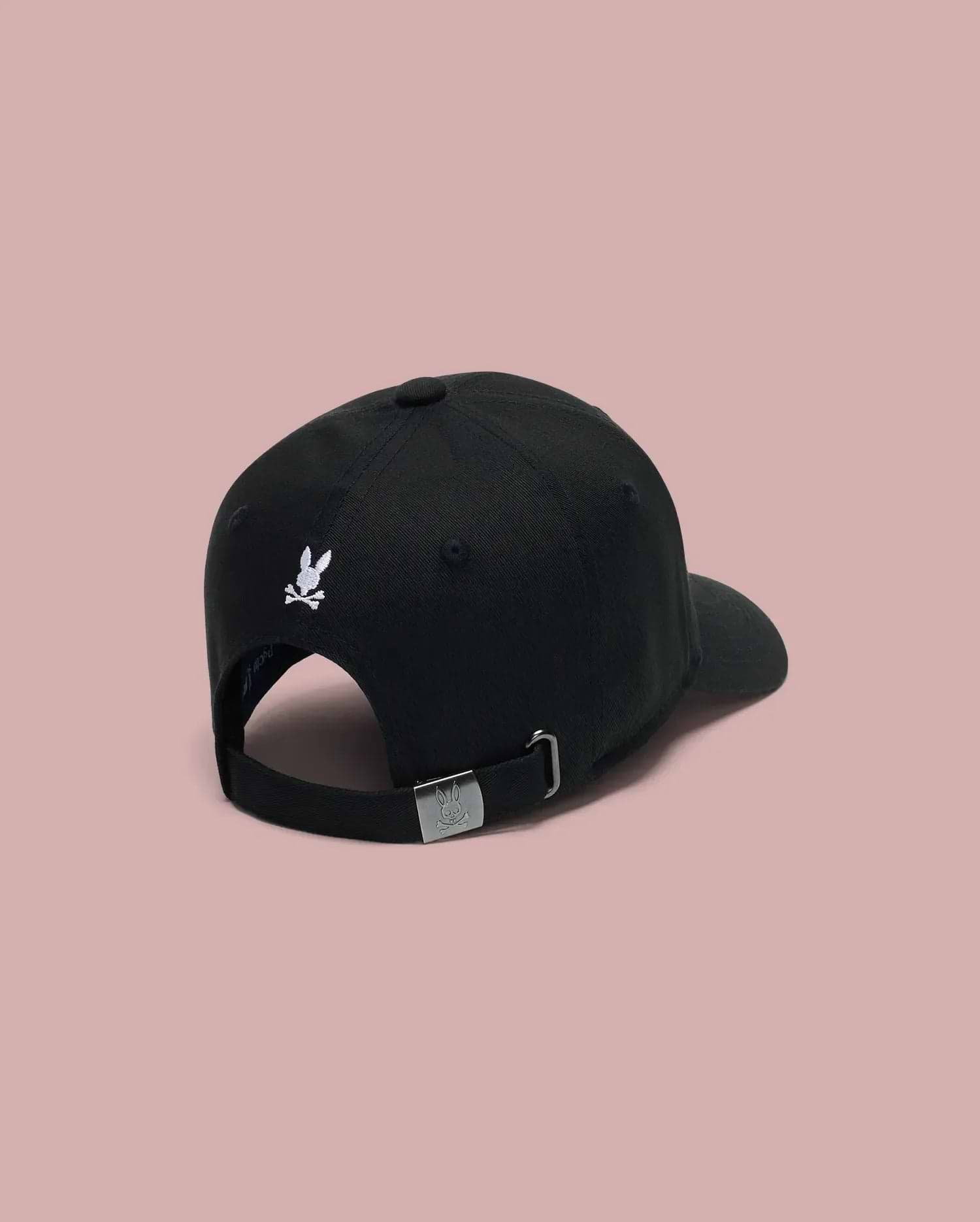 The Psycho Bunny MENS ARCHIVE BASEBALL CAP - MO1500002 is a 100% cotton black cap, shown from the back with an embroidered bunny logo above the opening and an adjustable strap with metal buckle, set against a pink background.
