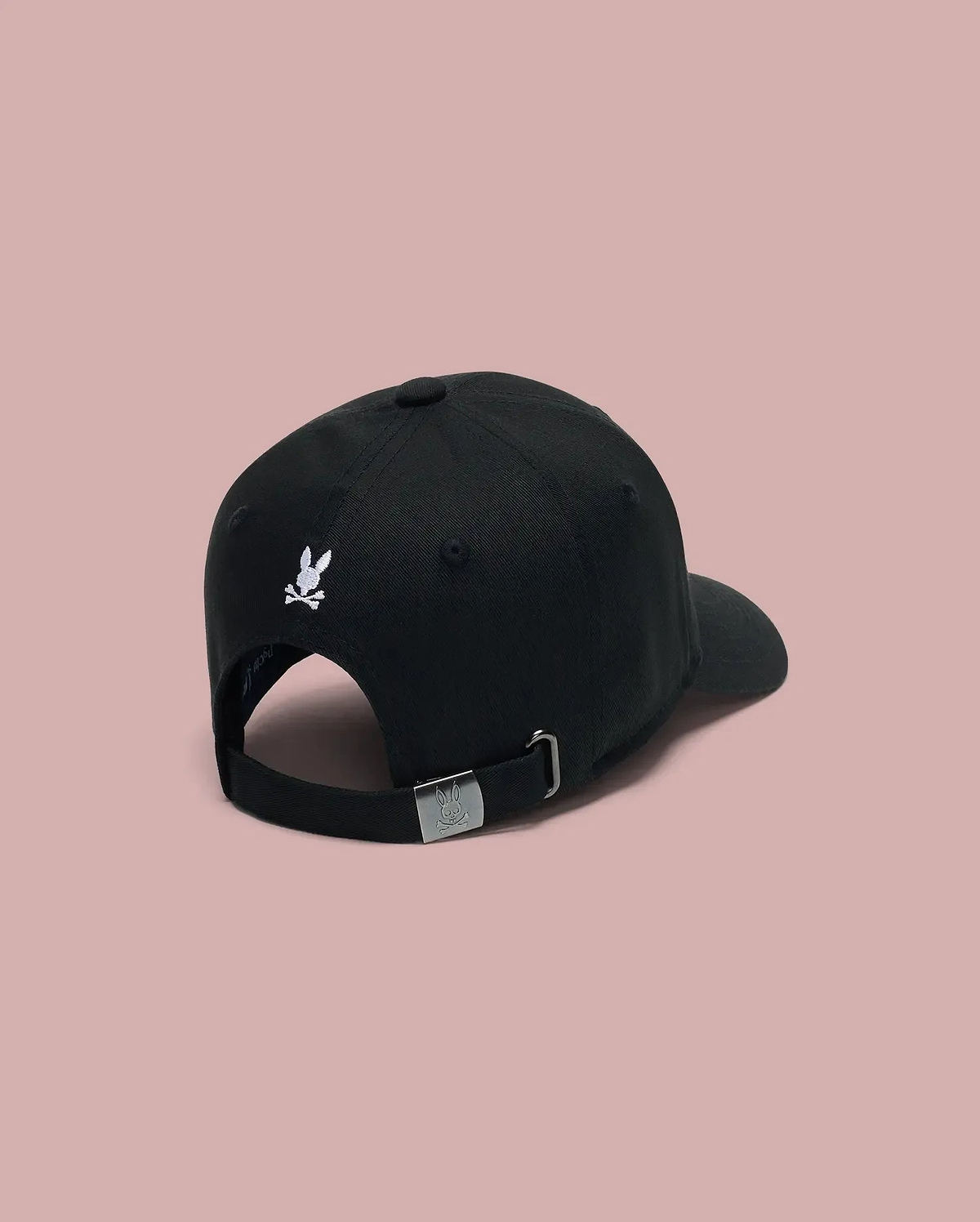 The Psycho Bunny MENS ARCHIVE BASEBALL CAP - MO1500002 is a 100% cotton black cap, shown from the back with an embroidered bunny logo above the opening and an adjustable strap with metal buckle, set against a pink background.