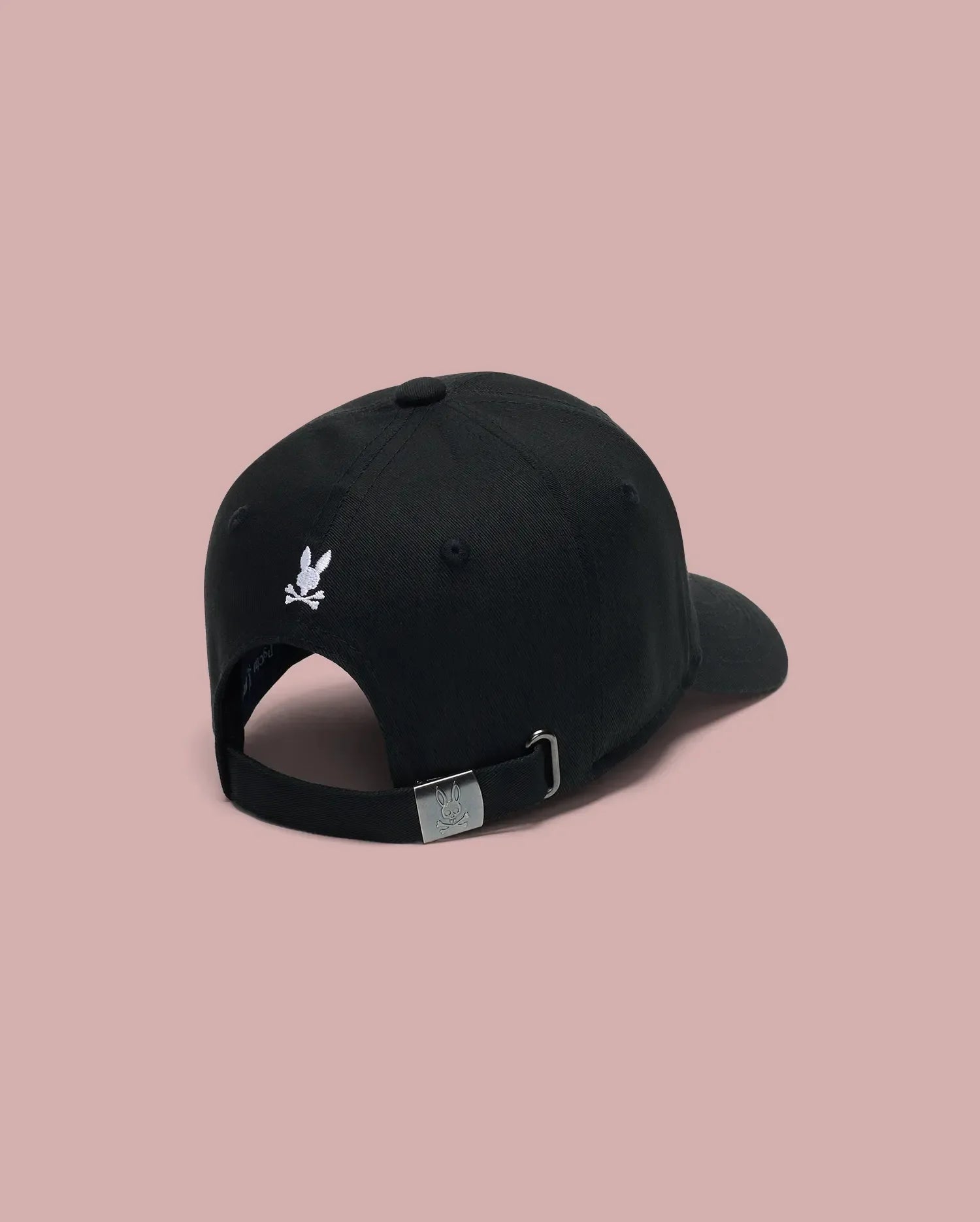 The Psycho Bunny MENS ARCHIVE BASEBALL CAP - MO1500002 is a 100% cotton black cap, shown from the back with an embroidered bunny logo above the opening and an adjustable strap with metal buckle, set against a pink background.