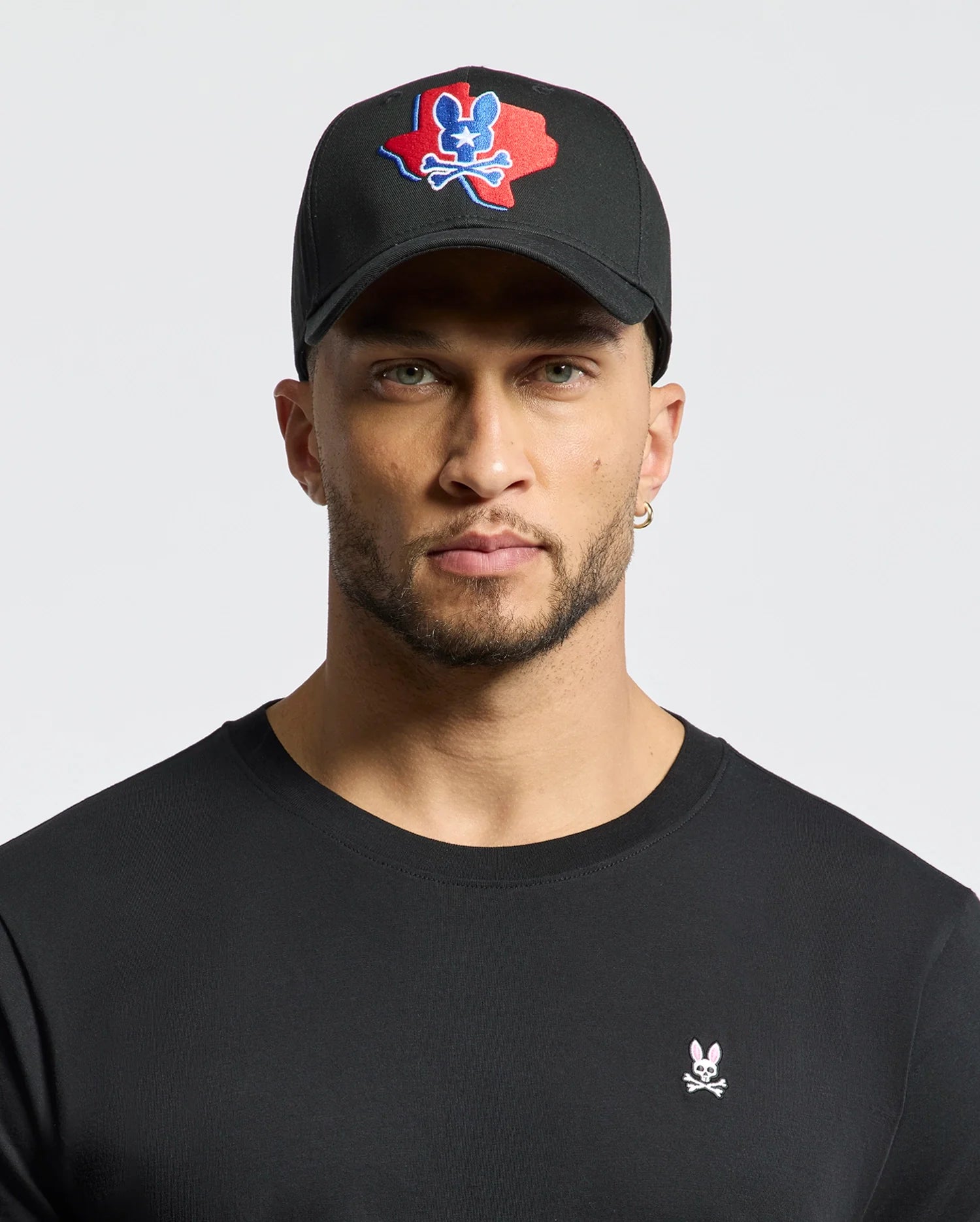 A man with light stubble wears a black Psycho Bunny MENS TEXAS BASEBALL CAP - MO1500014, featuring an embroidered red and blue skull design. He also sports a black shirt with a small woven skull and bunny logo against a plain white background. (1)