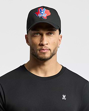 A man with light stubble wears a black Psycho Bunny MENS TEXAS BASEBALL CAP - MO1500014, featuring an embroidered red and blue skull design. He also sports a black shirt with a small woven skull and bunny logo against a plain white background. (1)