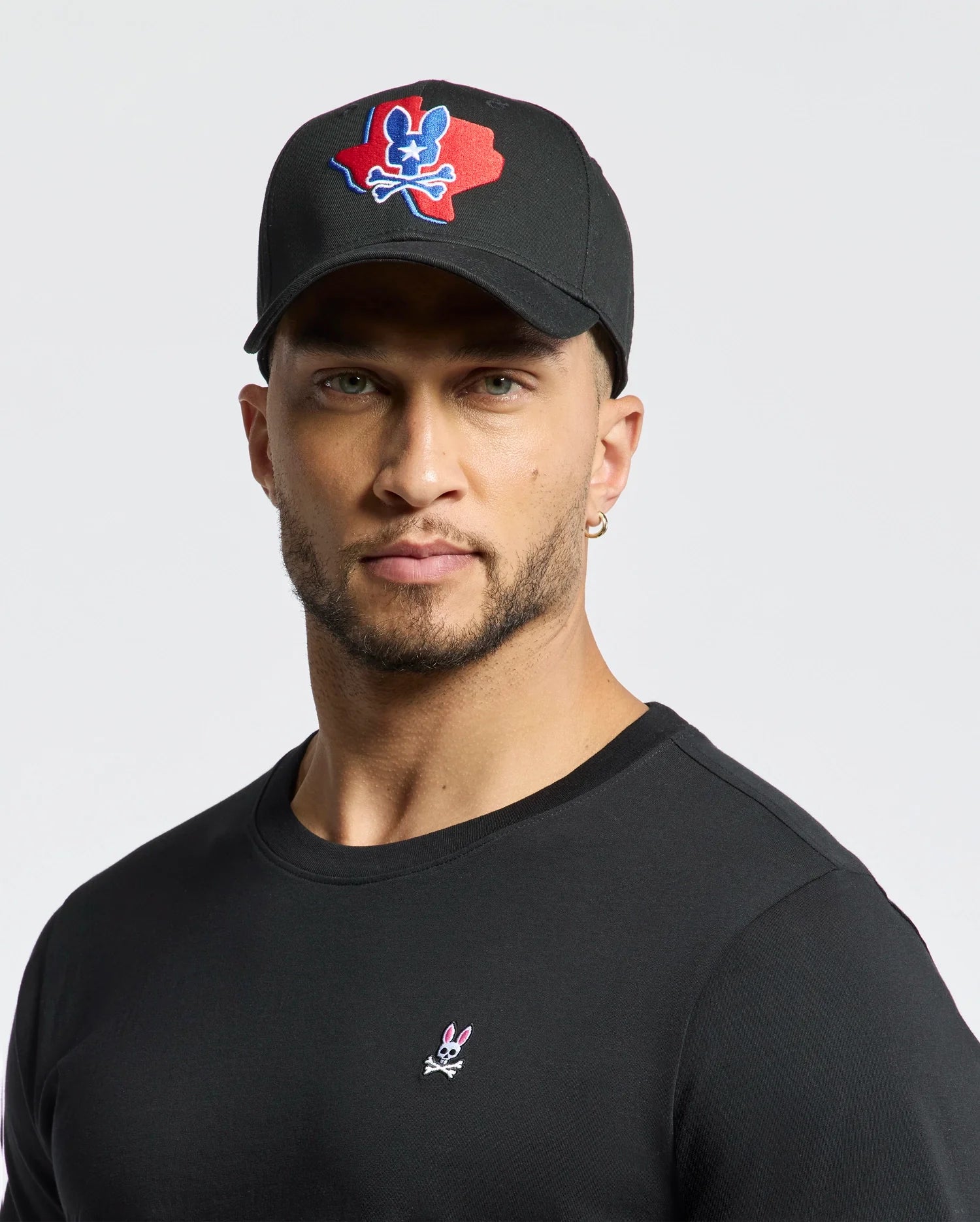 A man dons Psycho Bunny's MENS TEXAS BASEBALL CAP - MO1500014 with a colorful logo and a black cotton shirt embellished with a small design, set against a plain white background. (6)