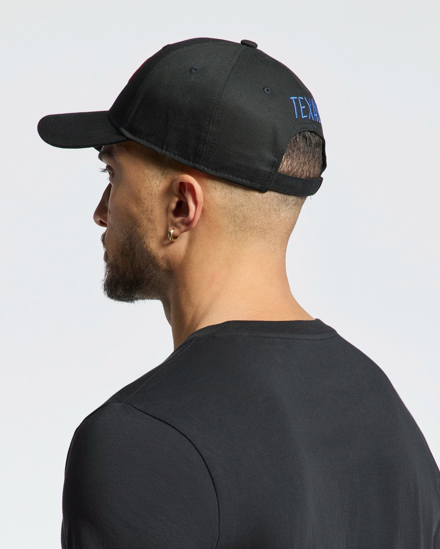 A bearded man is seen from the side and back, wearing a black cotton T-shirt and the MENS TEXAS BASEBALL CAP - MO1500014 by Psycho Bunny. The cap features blue woven text on the back. His head slightly turns left against a plain white background. (5)
