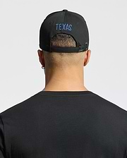 A person with short hair wears a Psycho Bunny "MENS TEXAS BASEBALL CAP - MO1500014," beautifully embroidered in blue on the back. They sport a black cotton t-shirt, an earring in the left ear, and stand against a light gray backdrop. (3)