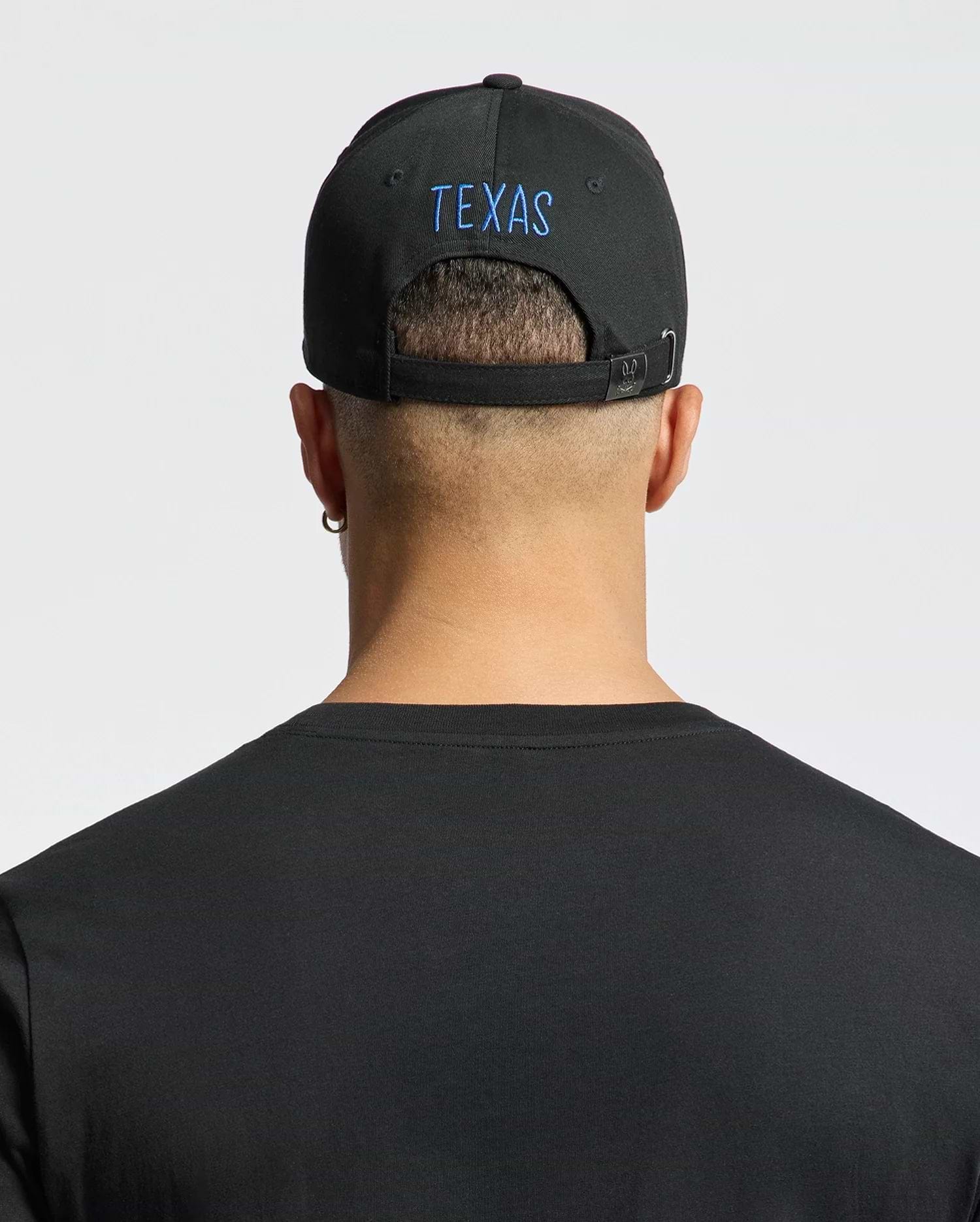A person with short hair wears a Psycho Bunny "MENS TEXAS BASEBALL CAP - MO1500014," beautifully embroidered in blue on the back. They sport a black cotton t-shirt, an earring in the left ear, and stand against a light gray backdrop. (3)