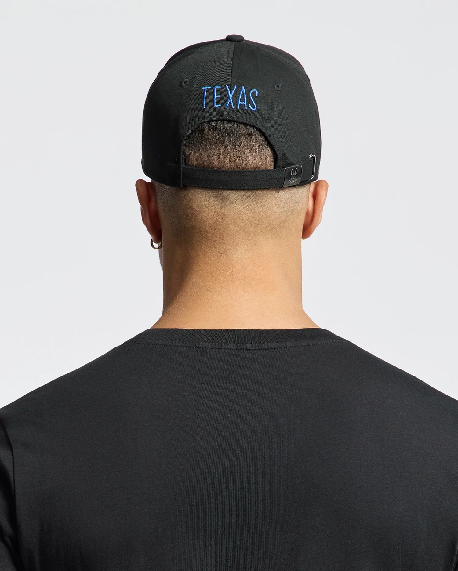 A person with short hair wears a Psycho Bunny "MENS TEXAS BASEBALL CAP - MO1500014," beautifully embroidered in blue on the back. They sport a black cotton t-shirt, an earring in the left ear, and stand against a light gray backdrop. (3)