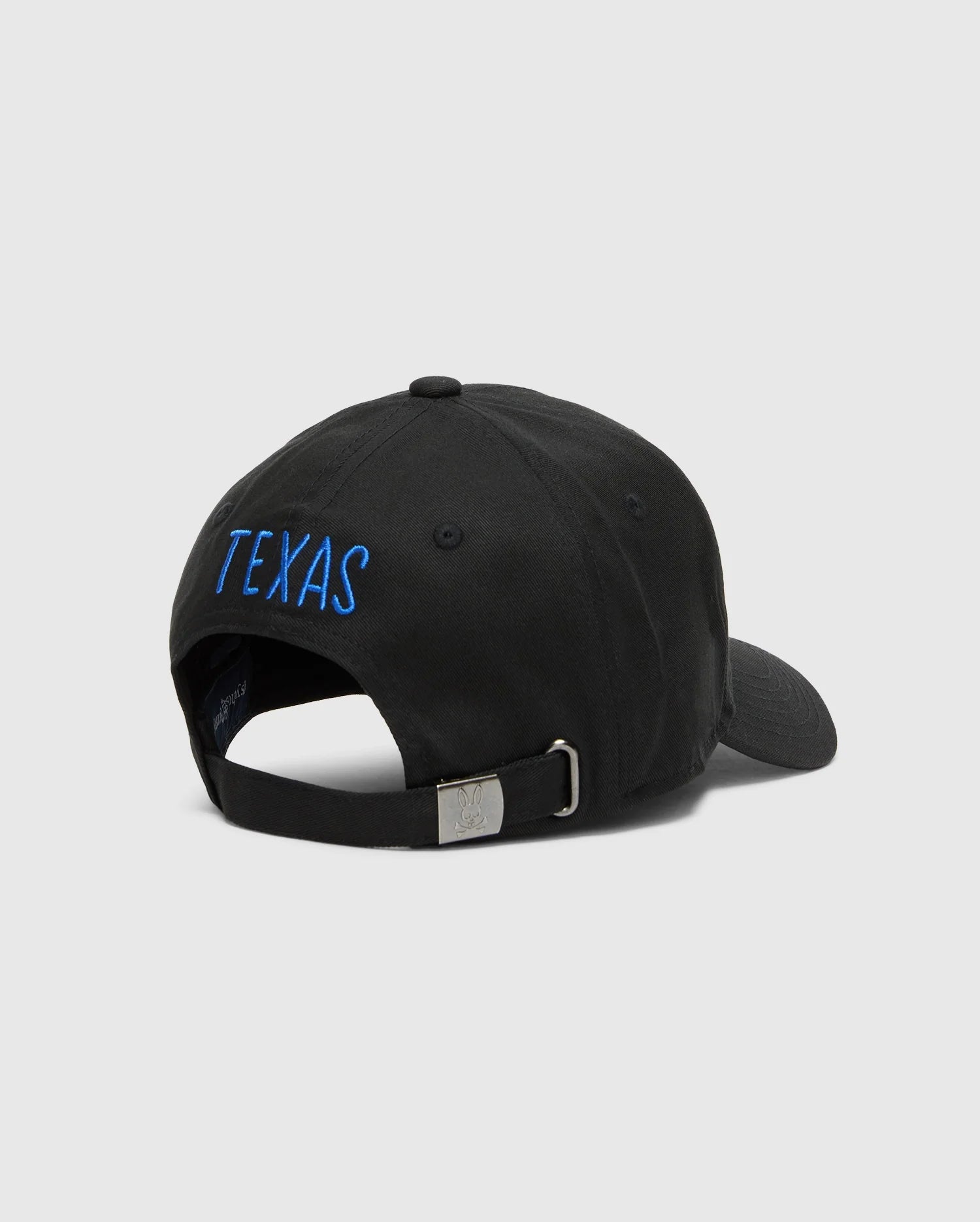 The Psycho Bunny MENS TEXAS BASEBALL CAP - MO1500014 features "TEXAS" embroidered in blue on a black cotton cap. It has a woven adjustable strap with a sleek metal buckle against a plain, light gray background. (4)