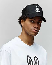A person wears a Psycho Bunny MENS RYDER TRUCKER CAP (MO1500030) in black cotton twill with a white skull and bunny logo, paired with a white tee featuring the matching bunny logo, standing against a plain light background.