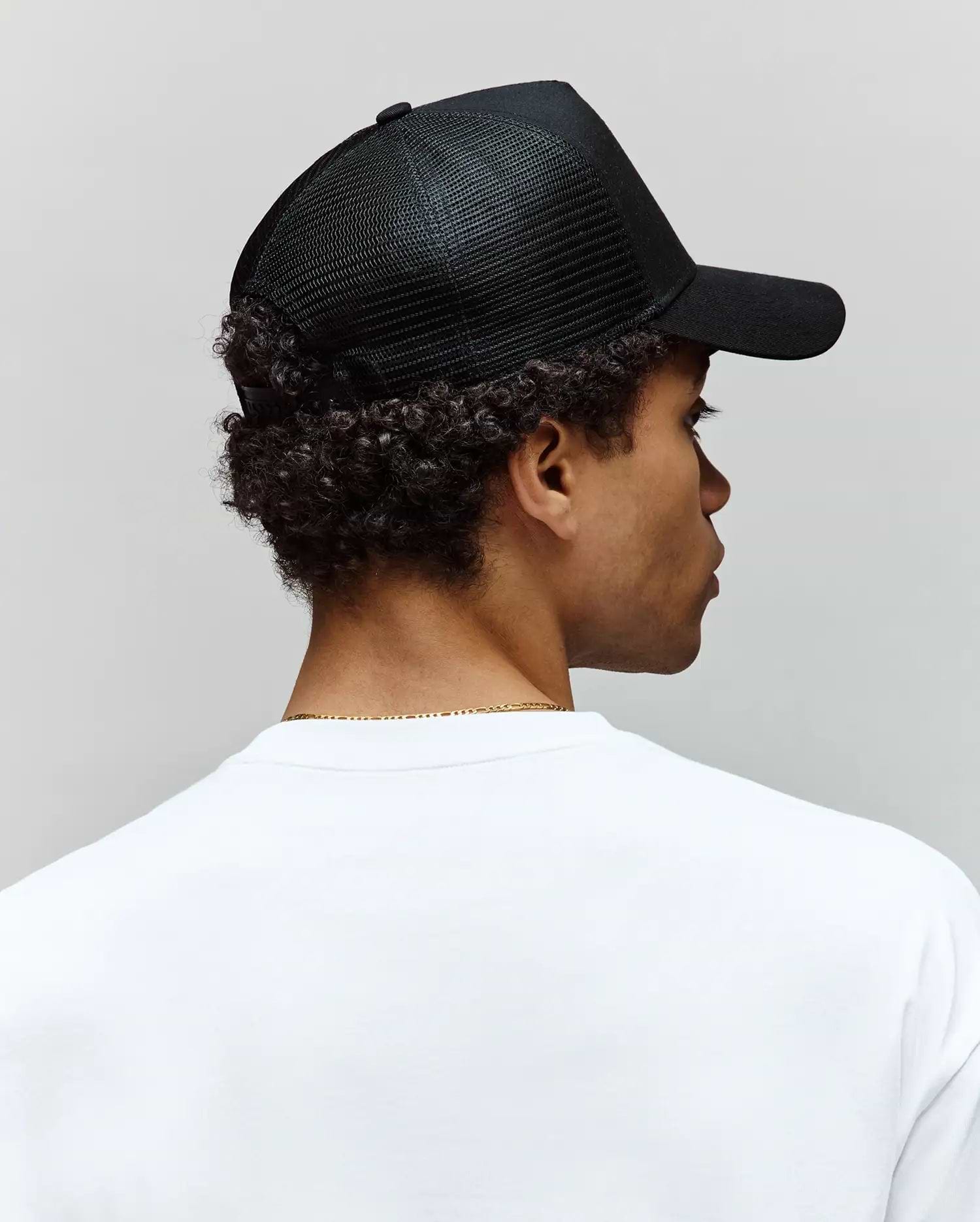 A person with short curly hair, seen from behind against a plain light background, wears a white T-shirt and a black Psycho Bunny MENS RYDER TRUCKER CAP - MO1500030 with an adjustable snapback.