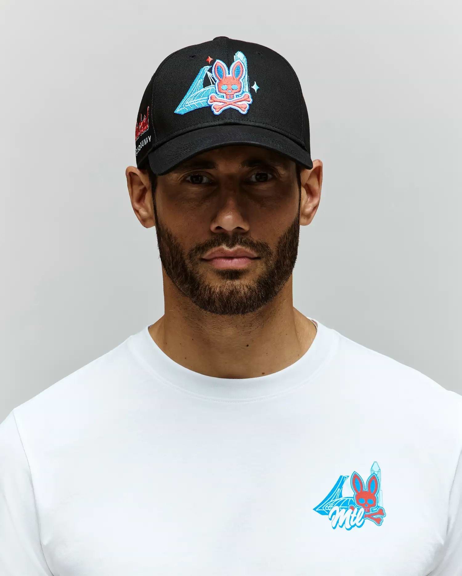 A bearded man stands against a light gray background wearing a Psycho Bunny MENS MONTREAL BASEBALL CAP - MO1500048 in black and a white 100% cotton t-shirt, both featuring an embroidered Bunny with blue triangle and pink accents.