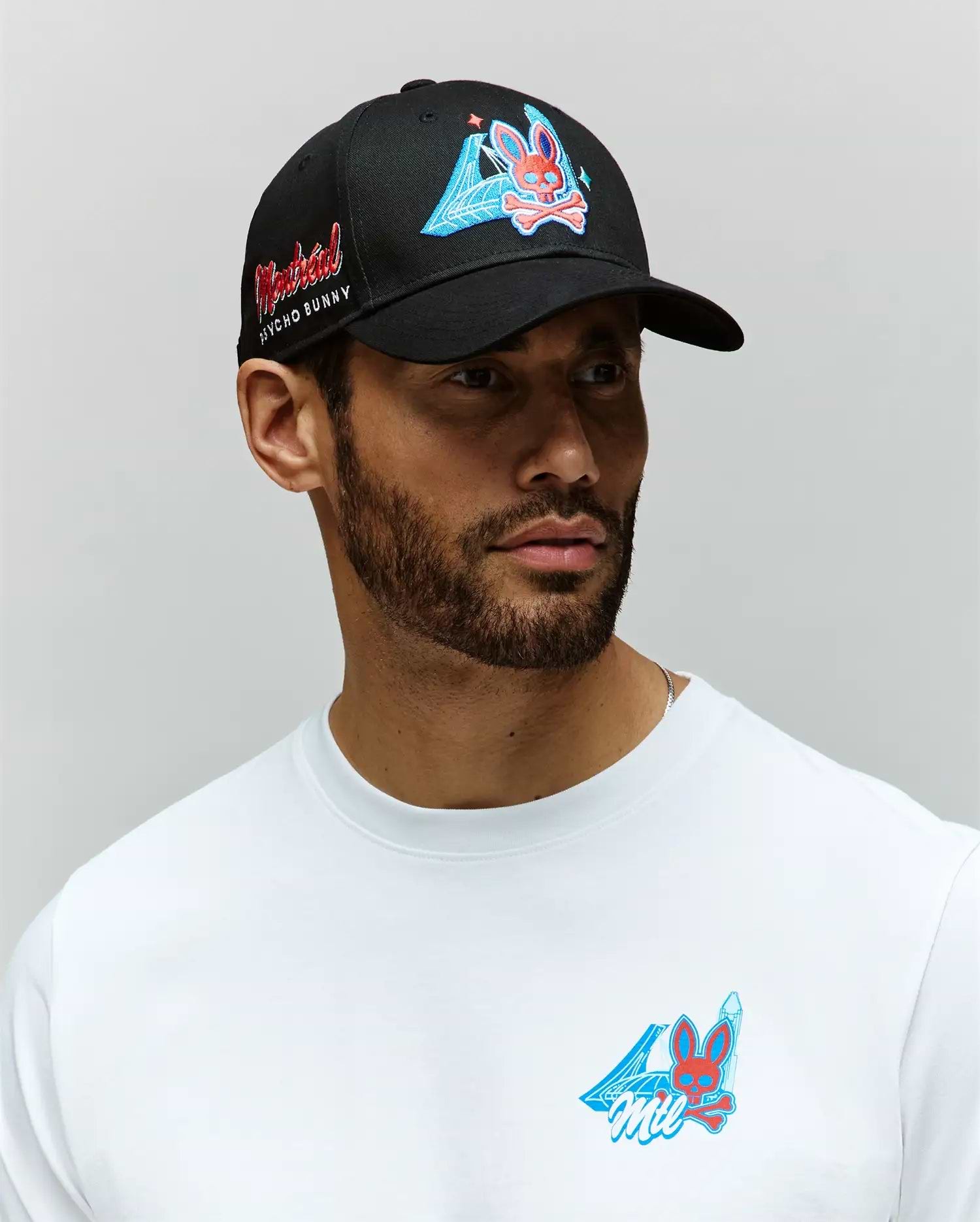 A man with a short beard wears a Psycho Bunny 100% cotton white shirt and the MENS MONTREAL BASEBALL CAP - MO1500048, both featuring colorful cartoon bunny graphics and blue geometric designs. The black cap has red embroidery on the side. Background is gray.