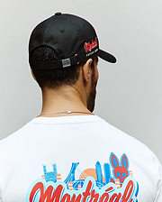 A man is seen from behind wearing a Psycho Bunny MENS MONTREAL BASEBALL CAP - MO1500048 in black with red side text, paired with a white 100% cotton tee featuring colorful city buildings and an embroidered Bunny graphic.