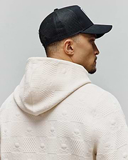 Ecru textured jacquard knit hoodie featuring an all-over pattern of alternating squares and Psycho Bunny skull logos, paired with a black Mens Adamo Trucker Cap with a white front logo.