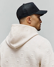 Ecru textured jacquard knit hoodie featuring an all-over pattern of alternating squares and Psycho Bunny skull logos, paired with a black Mens Adamo Trucker Cap with a white front logo.
