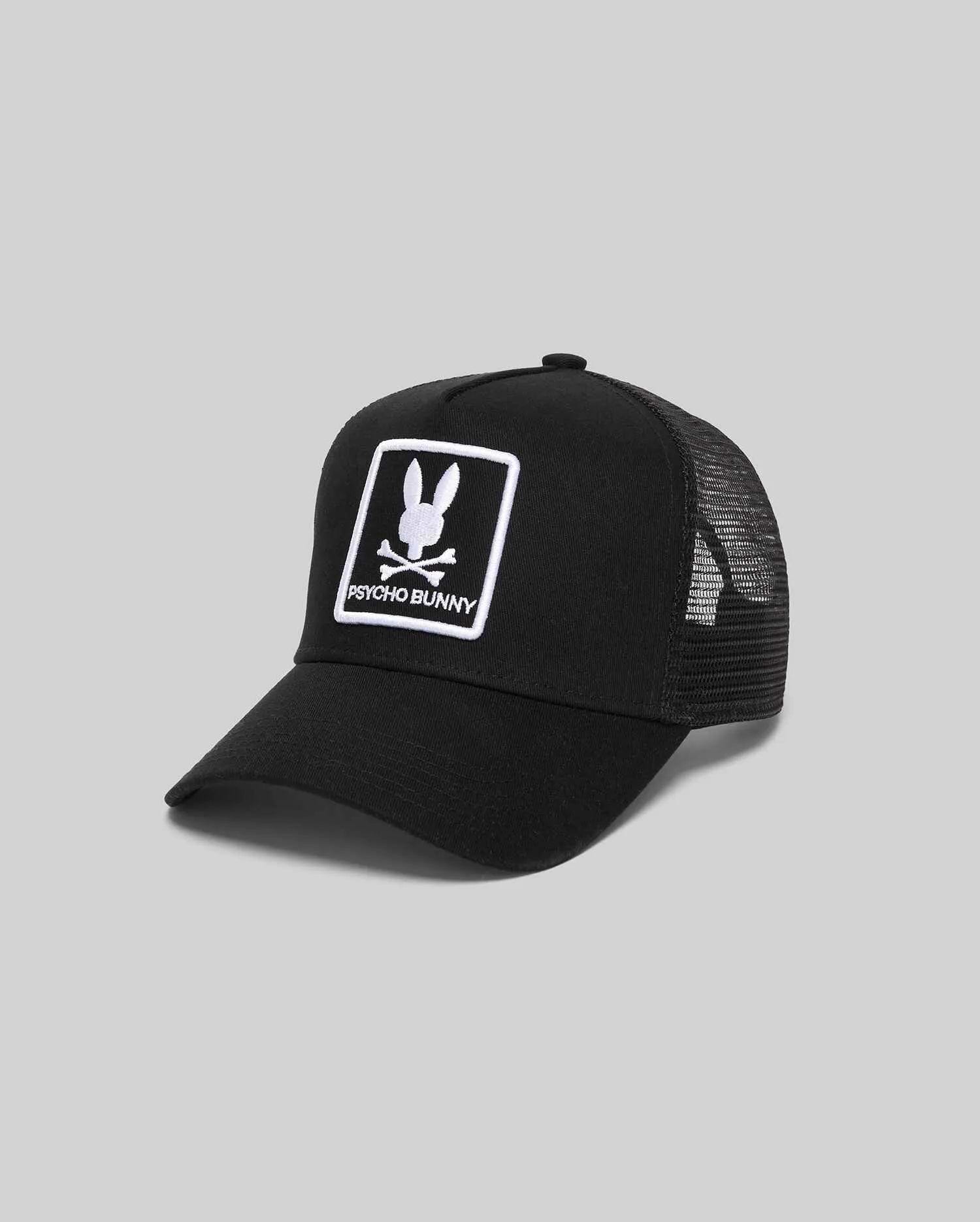 Black Mens Adamo Trucker Cap featuring a structured twill front and breathable mesh back. A prominent white embroidered Psycho Bunny skull logo patch with text adorns the front panel.