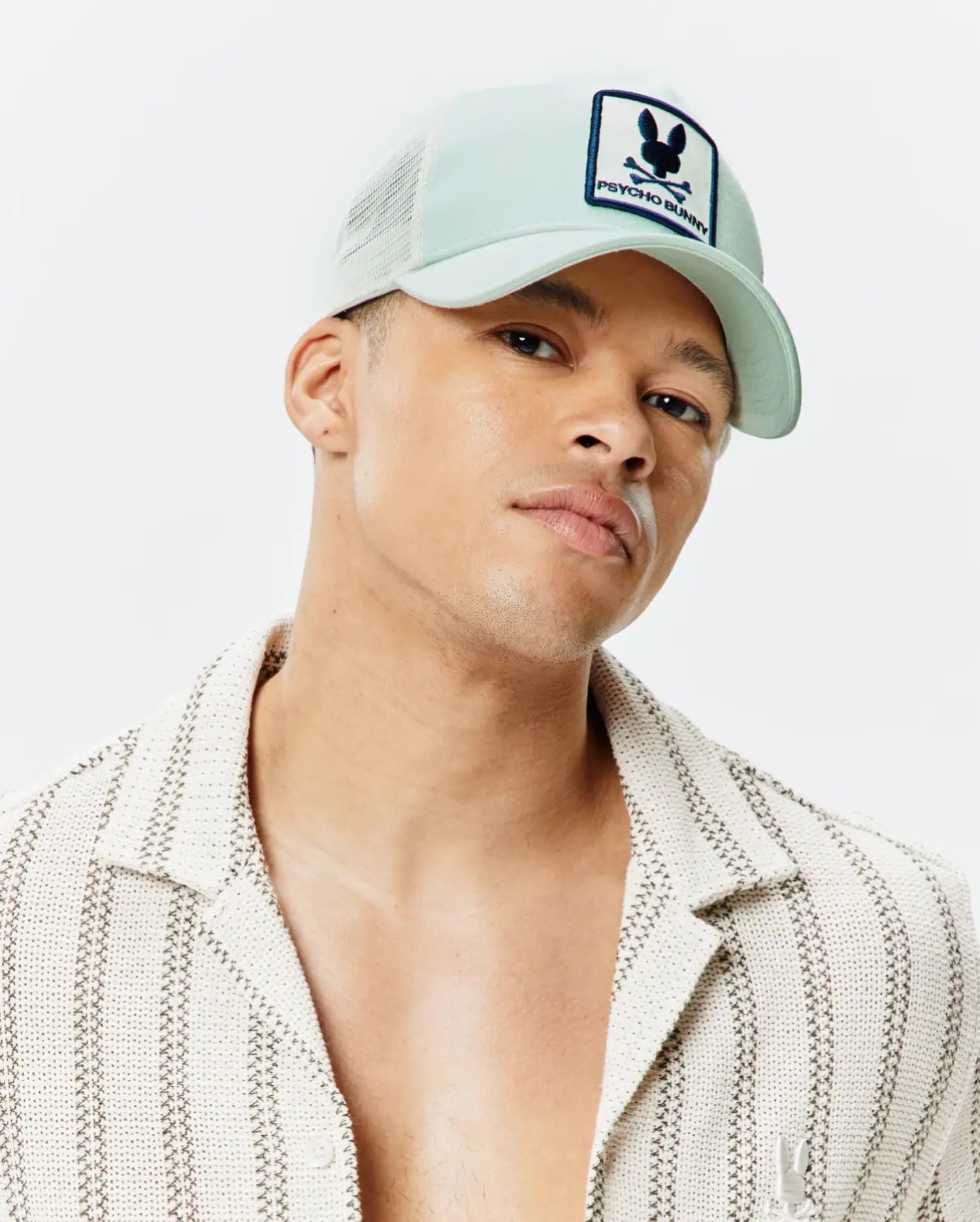 Mens Adamo Trucker Cap in light mint green with white mesh and a prominent Psycho Bunny skull logo patch, paired with a cream textured open shirt featuring dark vertical stripes and a subtle embroidered Psycho Bunny logo.