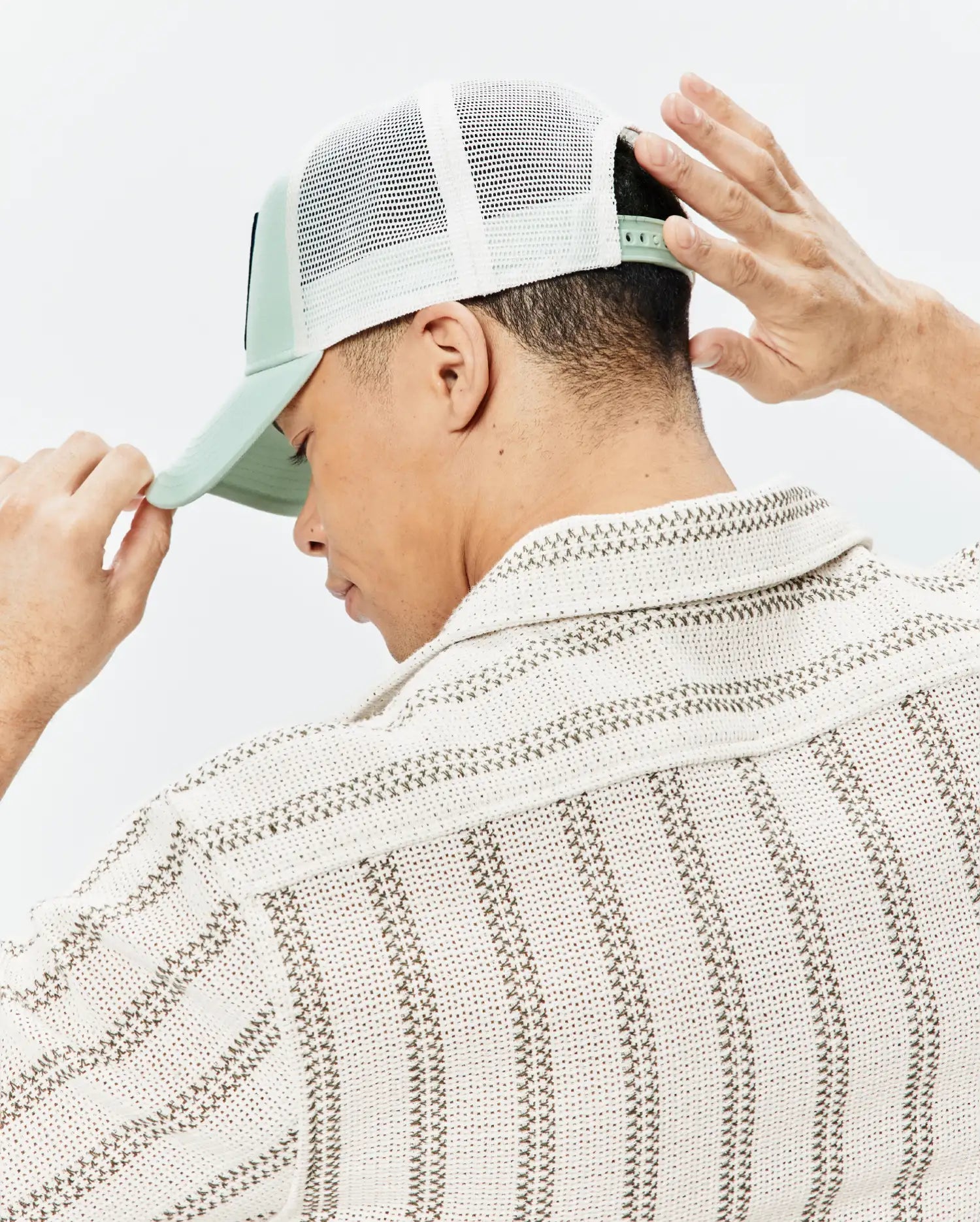 Psycho Bunny Mens Adamo Trucker Cap in sage green with front logo patch, white mesh back, and snapback, worn with a cream textured collared shirt featuring brown striped patterns.