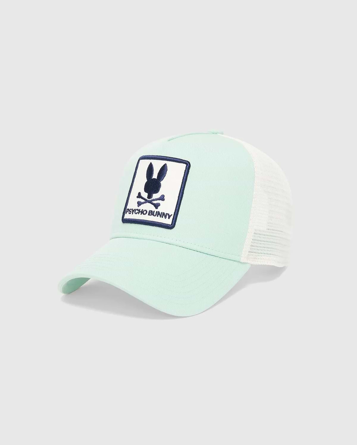 MENS ADAMO TRUCKER CAP in mint green cotton twill with white mesh back, featuring a large front embroidered patch with the navy blue Psycho Bunny skull logo and text on a white background.