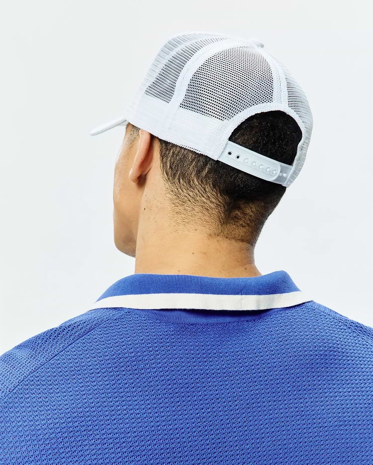 White Mens Adamo Trucker Cap with mesh back and snap closure, paired with a royal blue pique polo shirt featuring a blue and white striped collar.