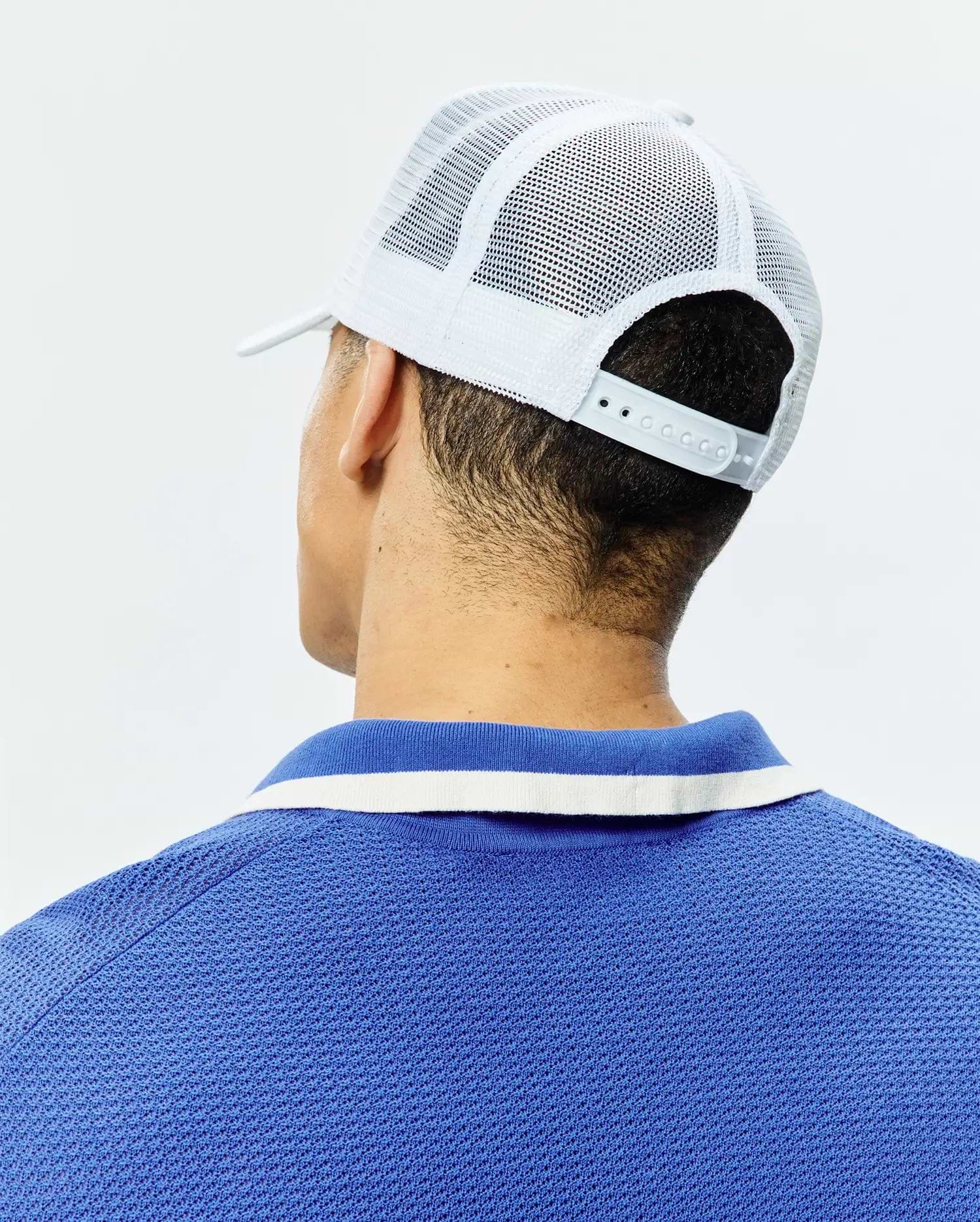 White Mens Adamo Trucker Cap with mesh back and snap closure, paired with a royal blue pique polo shirt featuring a blue and white striped collar.