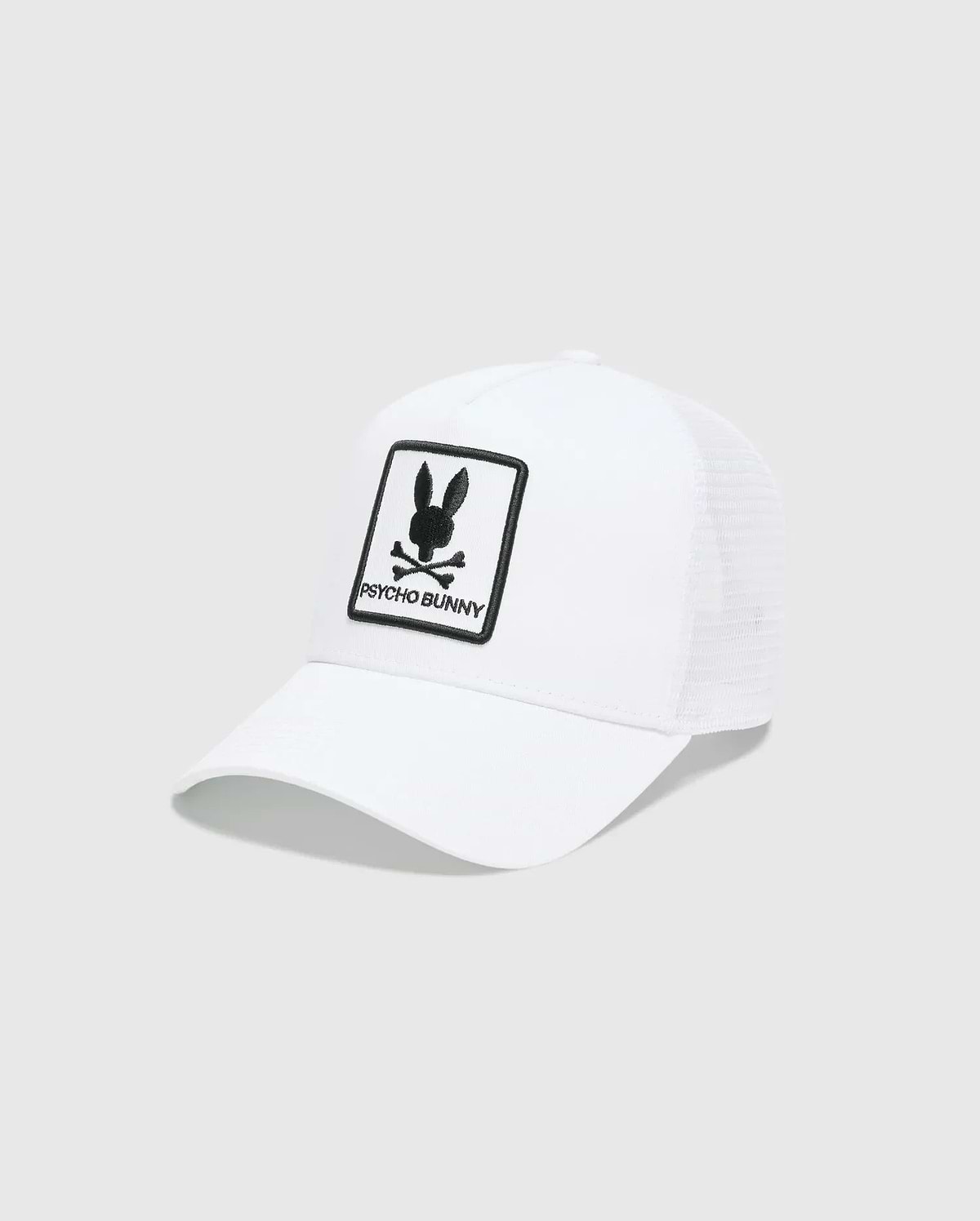 White Mens Adamo Trucker Cap featuring a large, centered black embroidered Psycho Bunny skull and crossbones logo patch with PSYCHO BUNNY text on the solid front panel and a breathable mesh back.