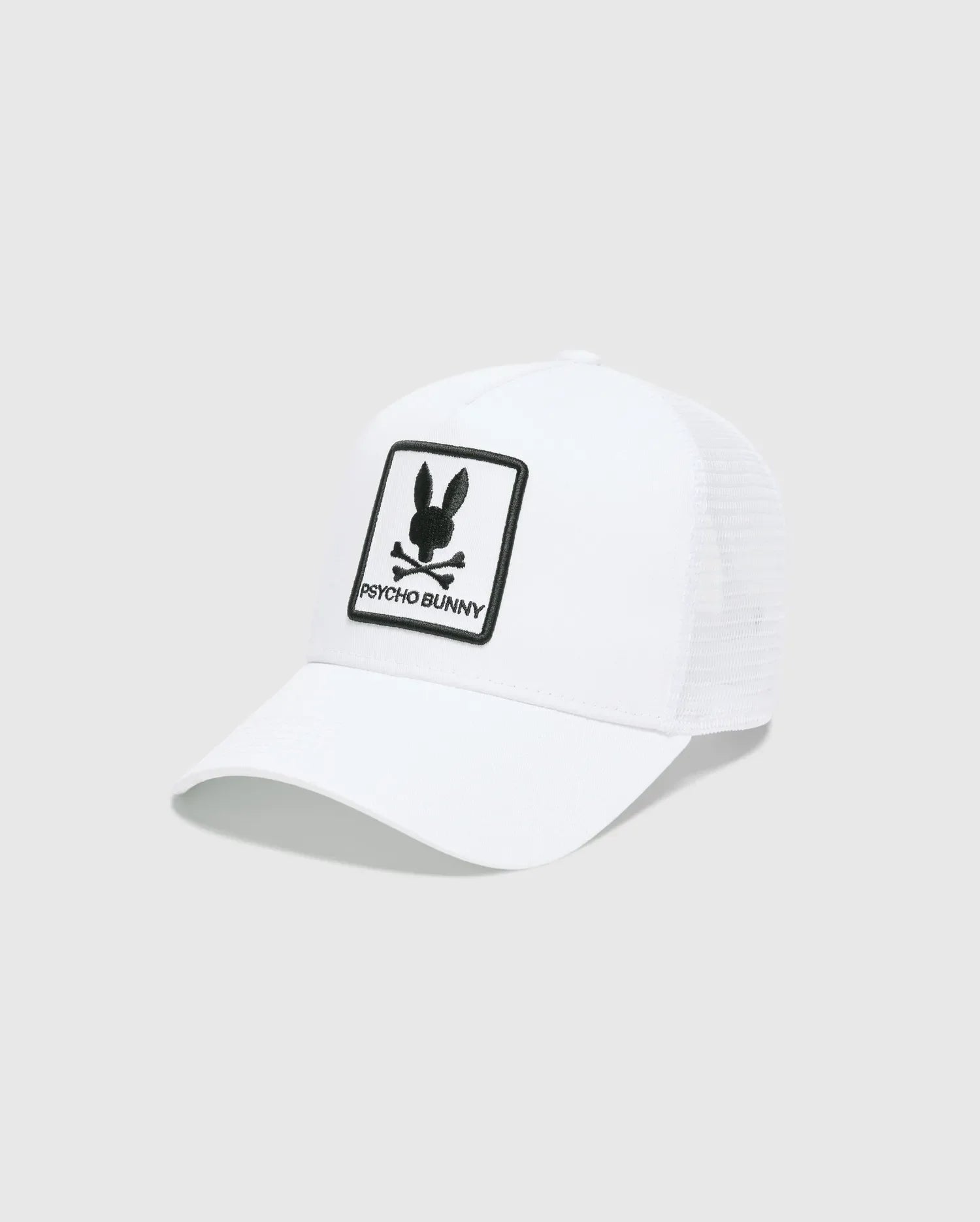 White Mens Adamo Trucker Cap featuring a large, centered black embroidered Psycho Bunny skull and crossbones logo patch with PSYCHO BUNNY text on the solid front panel and a breathable mesh back.